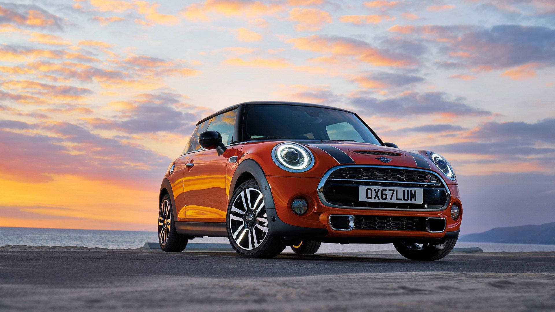 Mini Cooper 桌布 壁紙 — 一輛充滿活力的橙色MINI Cooper停泊在沿海公路上，天空呈現戲劇性的日落，帶有粉紅色和橙色的雲彩。
