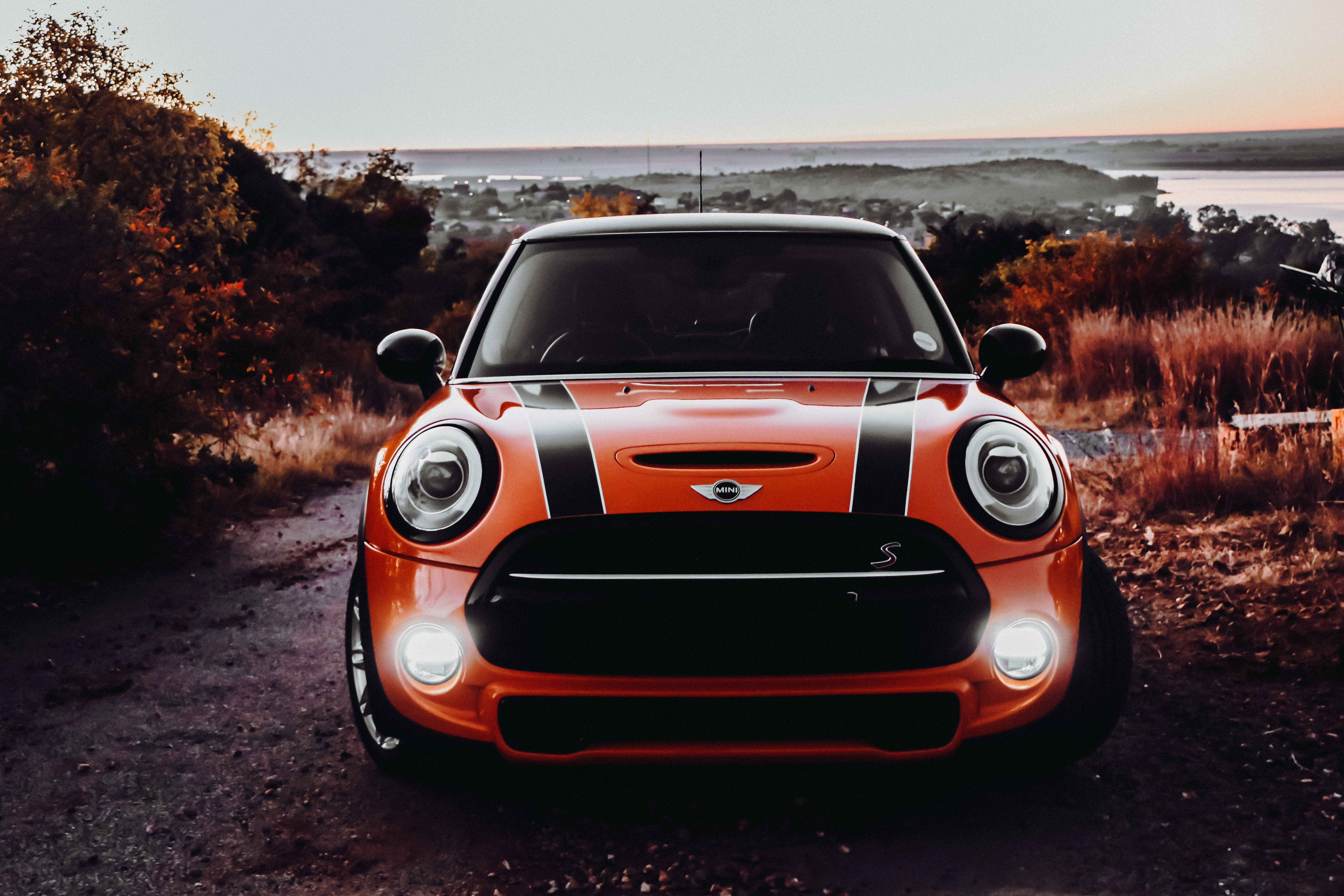Mini Cooper 桌布 壁紙 — 一輛充滿活力的橙色MINI Cooper S帶有黑色賽車條紋，在日落時分面向土路，車頭燈照亮秋天的樹葉和遠處的沿海小鎮。