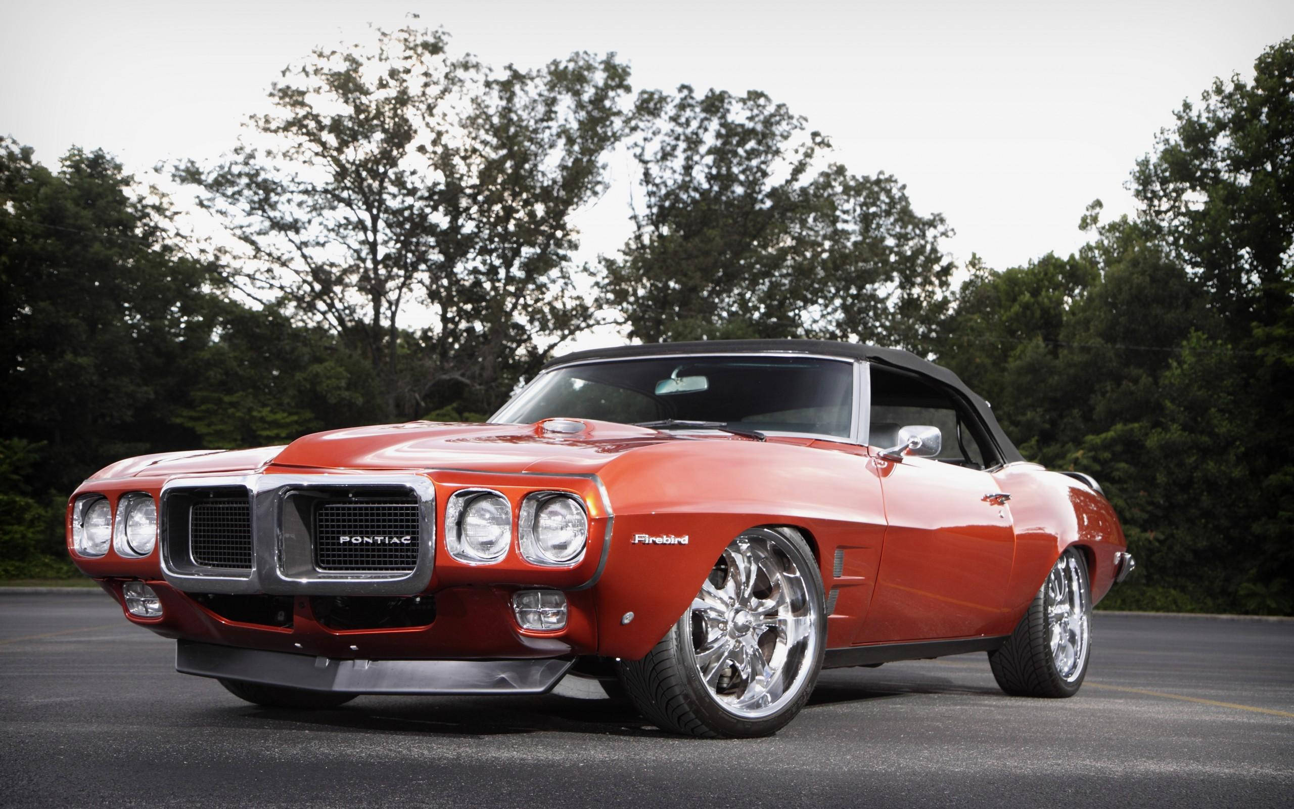 龐蒂克 桌布 壁紙 — 一輛充滿活力的橙色Pontiac Firebird敞篷車，配備鍍鉻輪圈，停在柏油路上，周圍綠樹成蔭。
