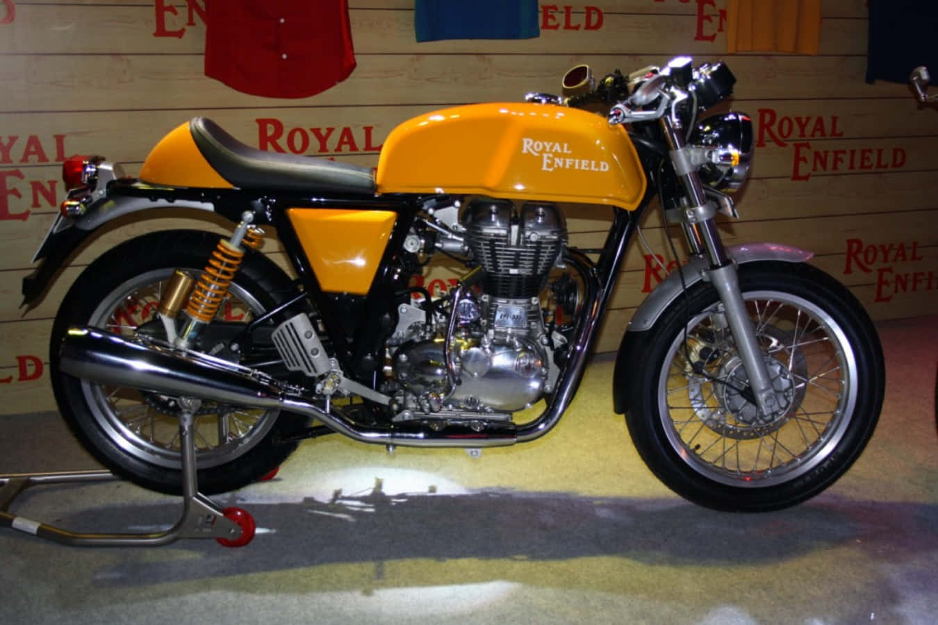 機車 桌布 壁紙 — 一輛充滿活力的橙色Royal Enfield摩托車在品牌展示間中，在聚光燈下閃閃發光，展示間有木牆和掛著的襯衫。