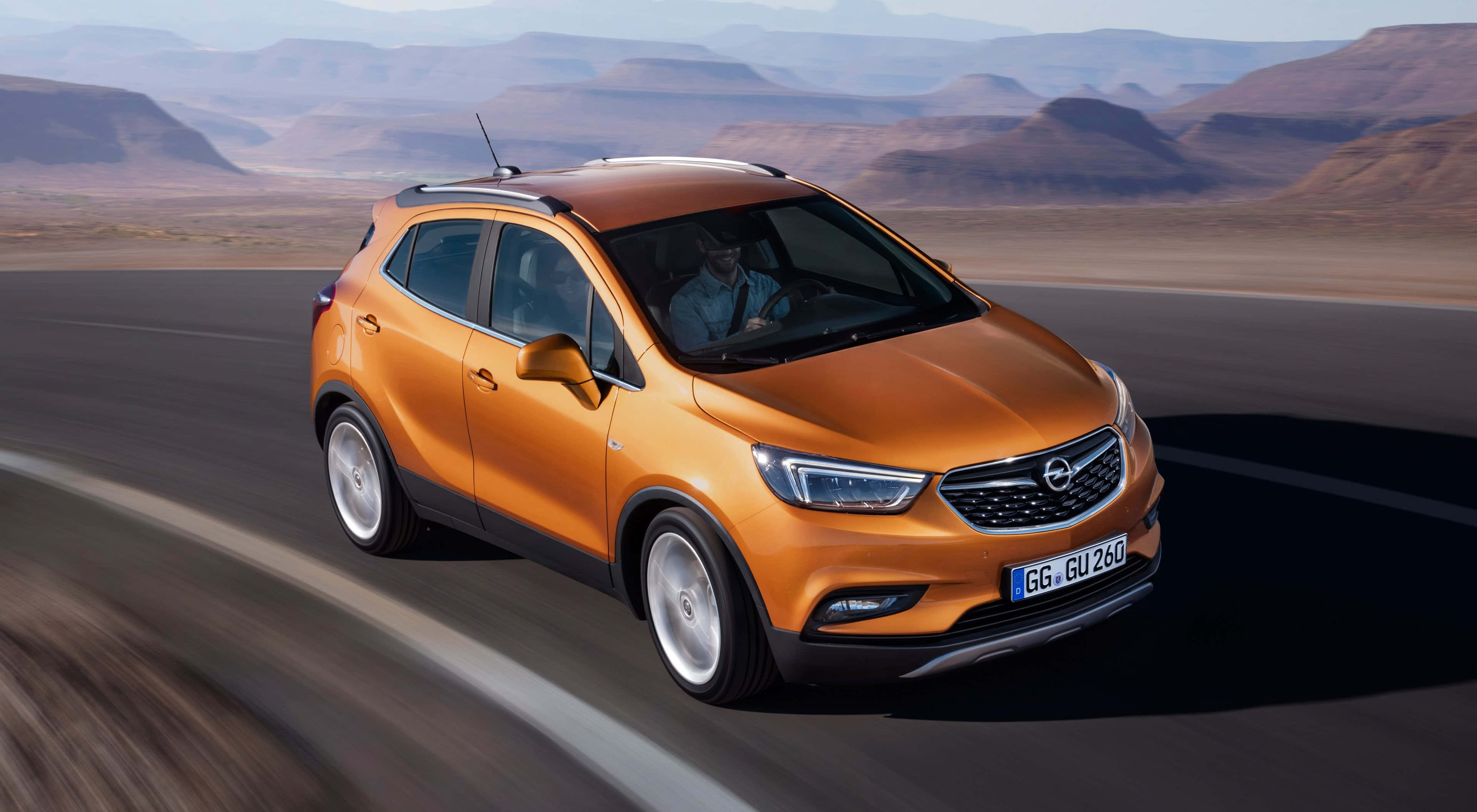 Opel Mokka 桌布 壁紙 — 一輛充滿活力的橙色SUV在沙漠公路上疾馳，遠處是朦朧的崎嶇山脈。