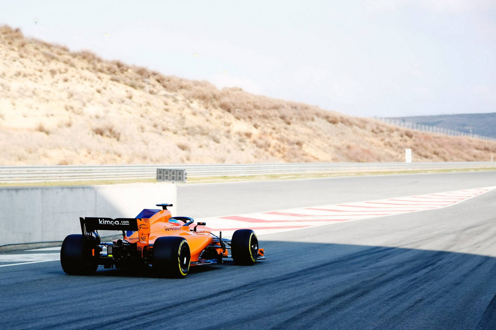 F1 桌布 壁紙 — 一輛充滿活力的橙色一級方程式賽車沿著陽光明媚的賽道飛馳，賽道兩旁是乾燥的山丘和安全護欄。