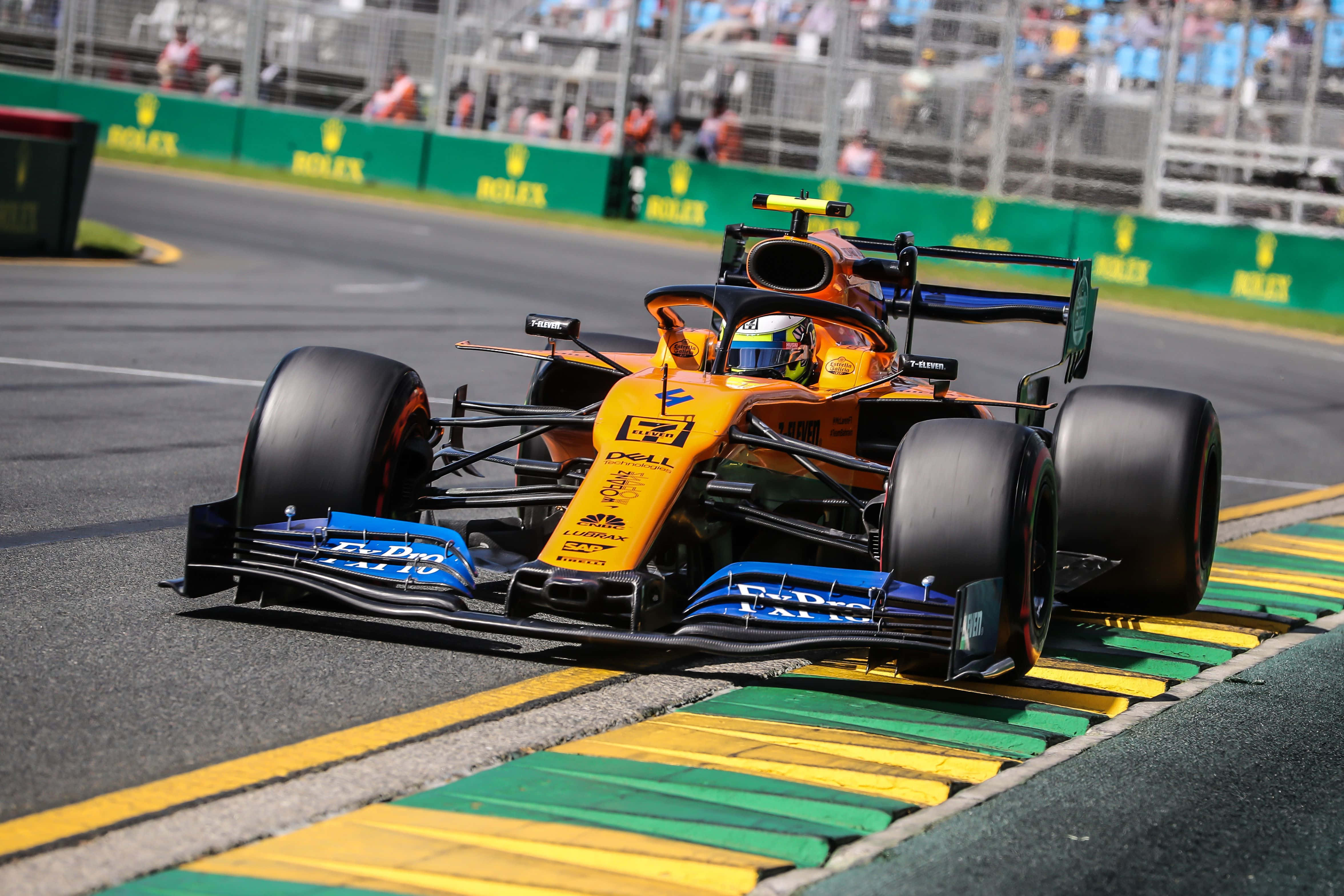lando norris 桌布 壁紙 — 一輛充滿活力的橙色和藍色一級方程式賽車在明亮的日光下，沿著由綠色和黃色路緣石環繞的賽道疾馳。