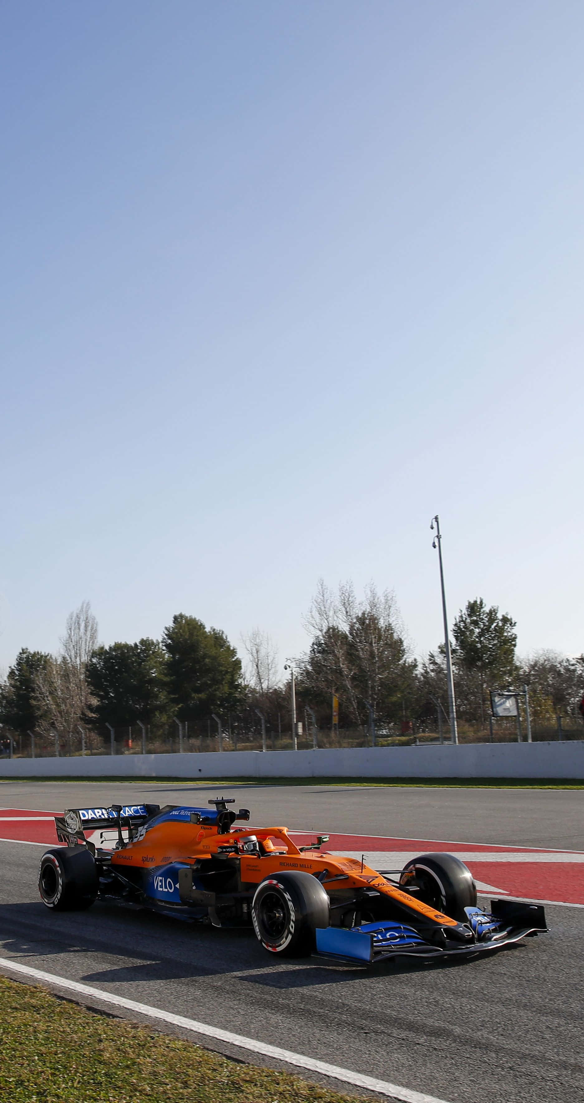 Formula 1 iPhone 桌布 壁紙 — 一輛充滿活力的橙色和藍色一級方程式賽車沿著陽光明媚的賽道飛馳，賽道兩旁是紅色路緣和光禿禿的樹木。