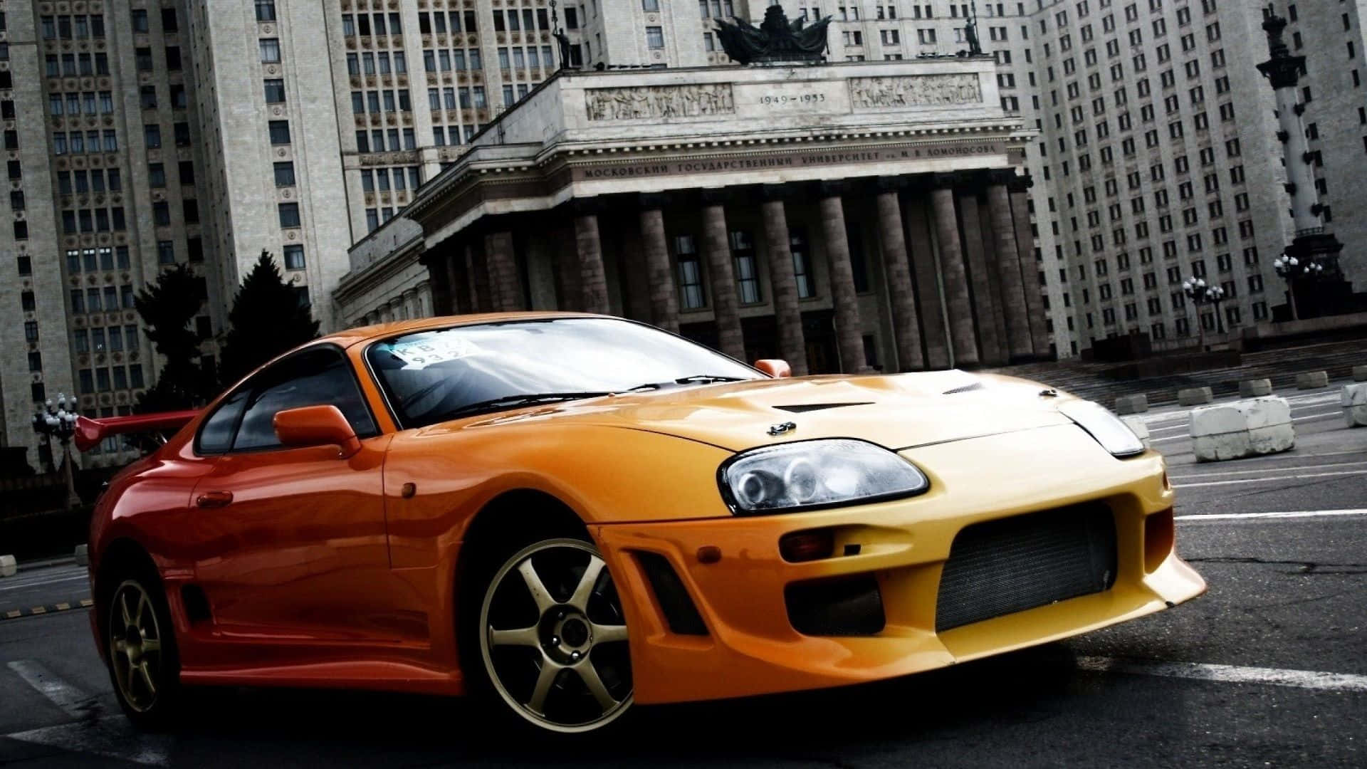 Aesthetic Car 桌布 壁紙 — 一輛充滿活力的橙色和黃色豐田Supra跑車停放在宏偉古典建築前的城市街道上。