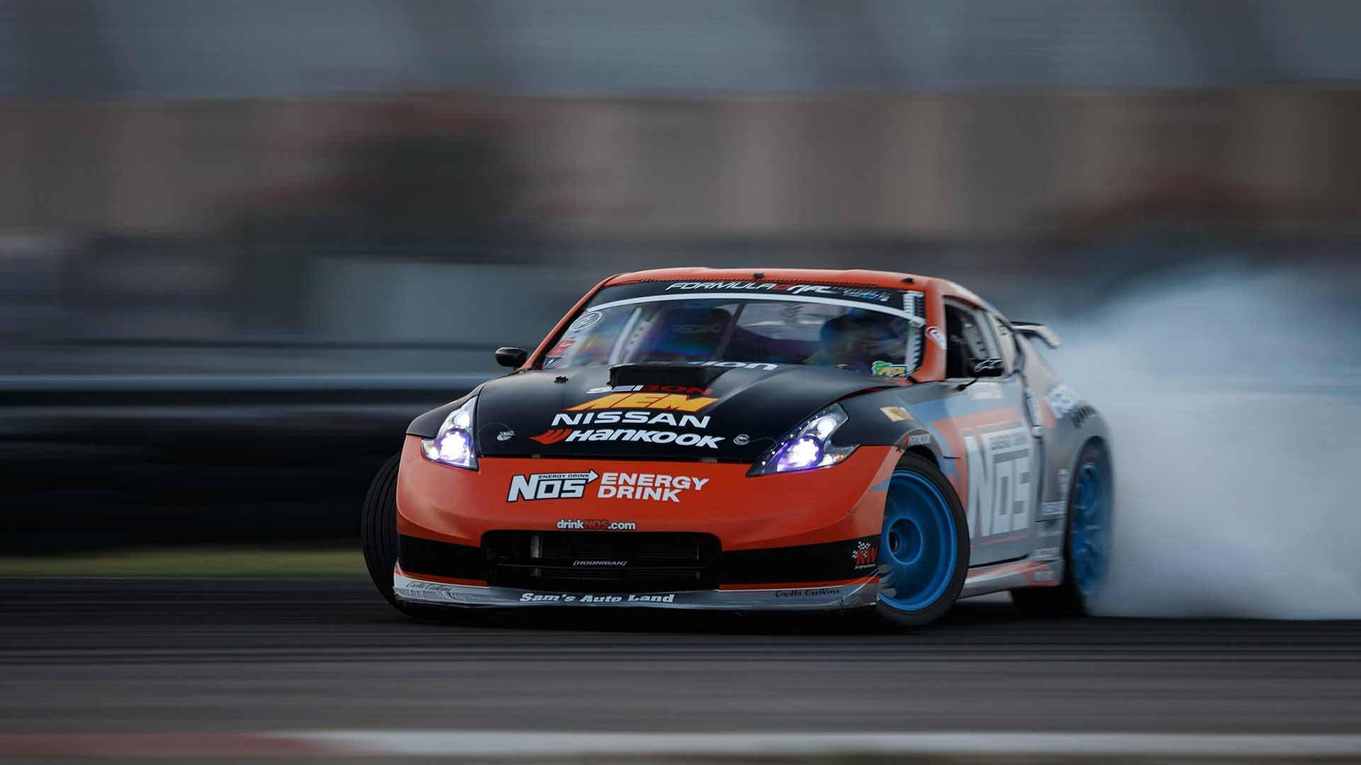 Nissan 350z 桌布 壁紙 — 一輛充滿活力的橙色和黑色甩尾車，配備藍色輪胎，在賽道彎角處激進地冒煙。