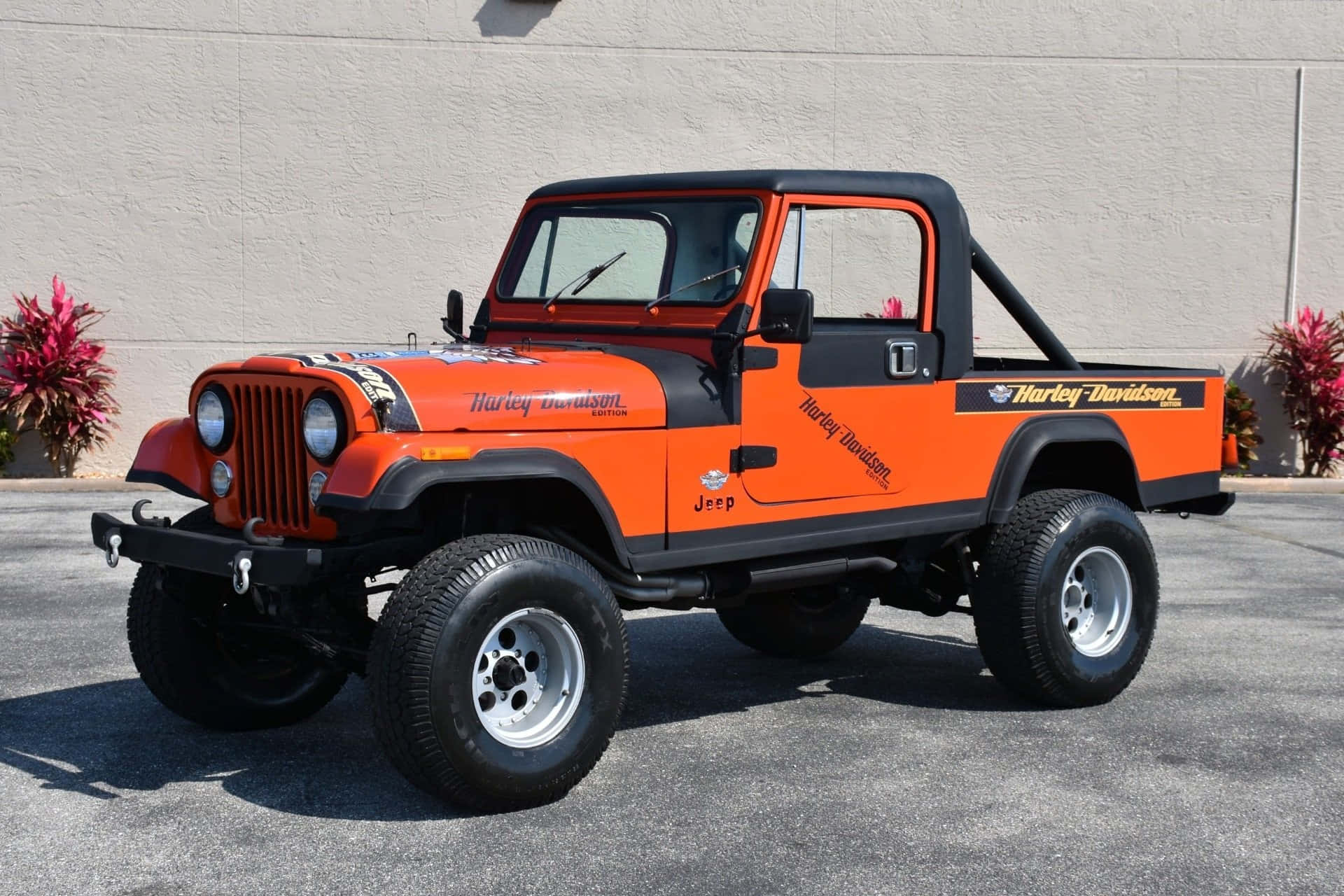 Jeep Cj 桌布 壁紙 — 一輛充滿活力的橙色哈雷戴維森品牌Jeep Wrangler，帶有黑色點綴，停在柏油路上，旁邊是紅葉植物。