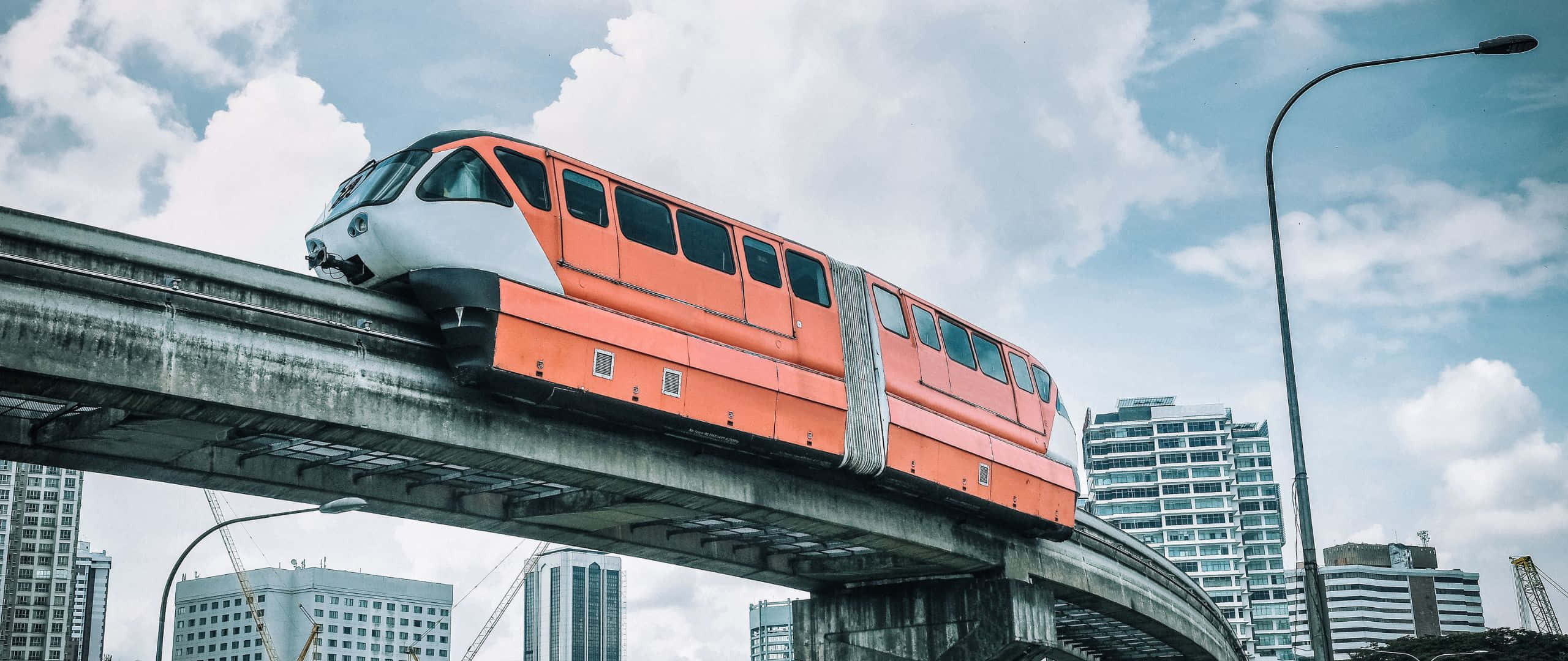 現代城市摩天大樓和多雲天空下，高架軌道上的橙色單軌列車。 — 2560x1080 動態桌布 壁紙