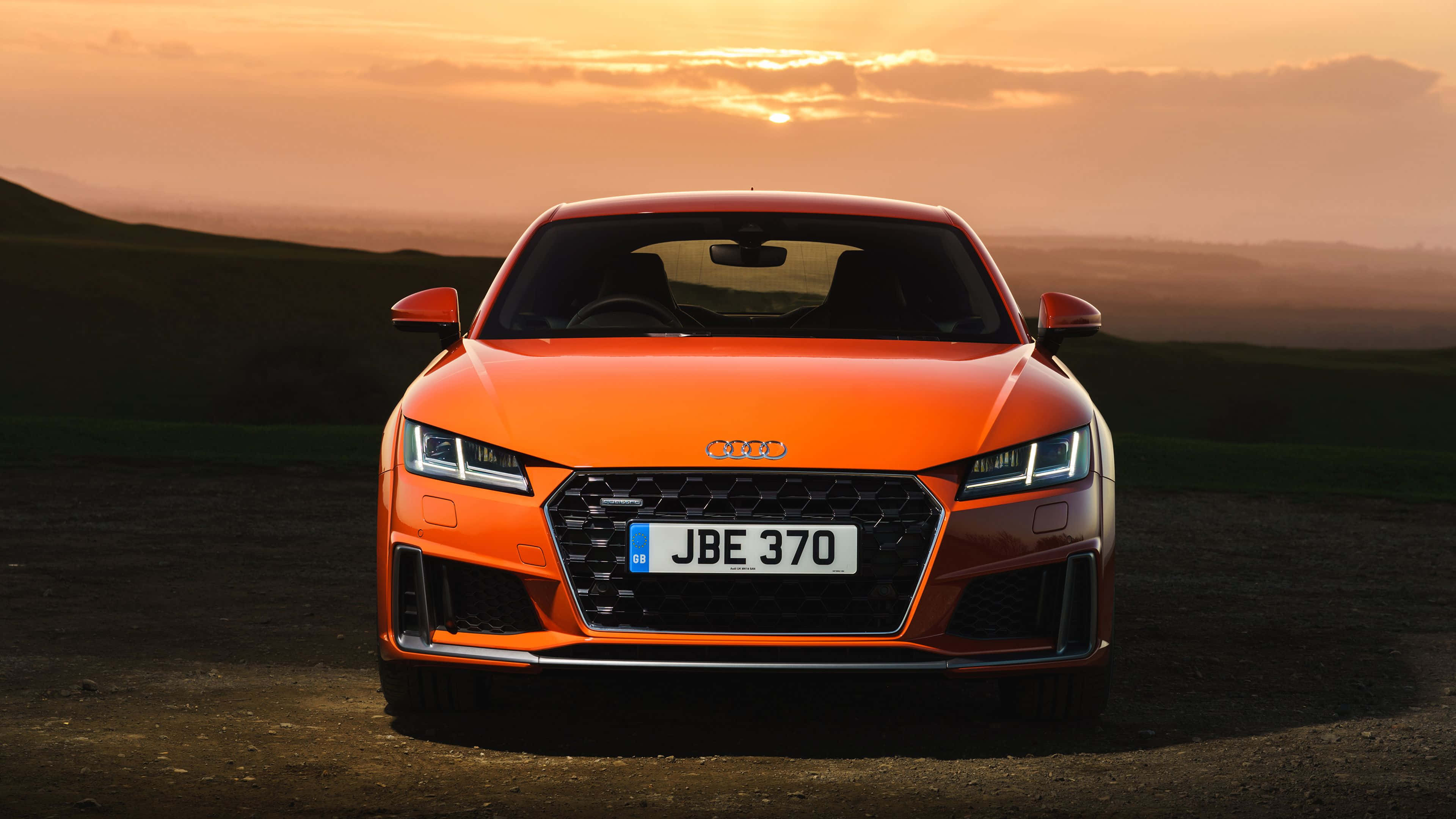 Audi Tt 桌布 壁紙 — 一輛充滿活力的橙色奧迪TT跑車在日落時分的泥路上向前行駛，背景是起伏的丘陵。