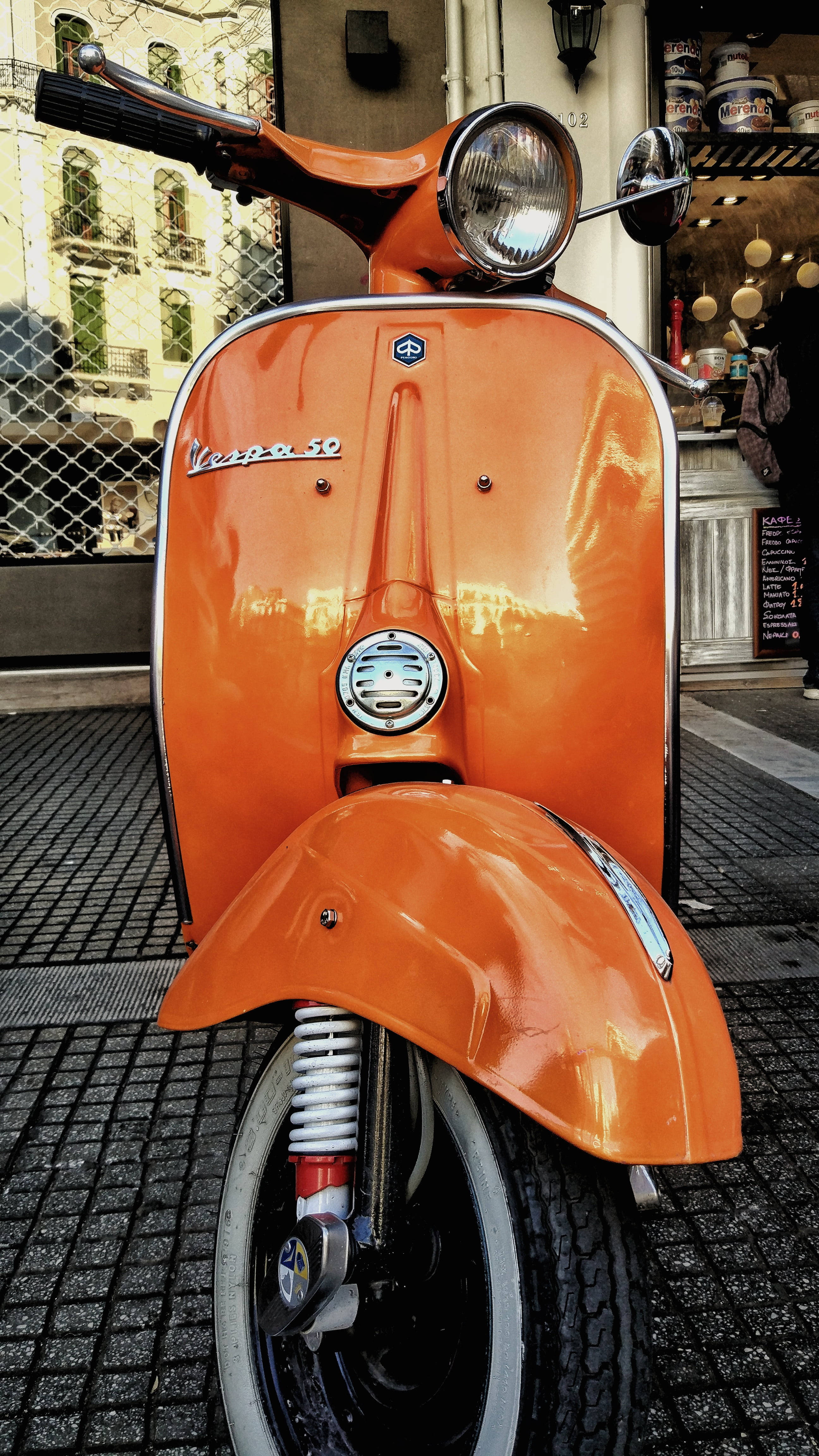 Retro Aesthetic 桌布 壁紙 — 一輛充滿活力的橙色復古Vespa 50踏板車在咖啡館旁的鵝卵石街道上，在日光下閃閃發光。