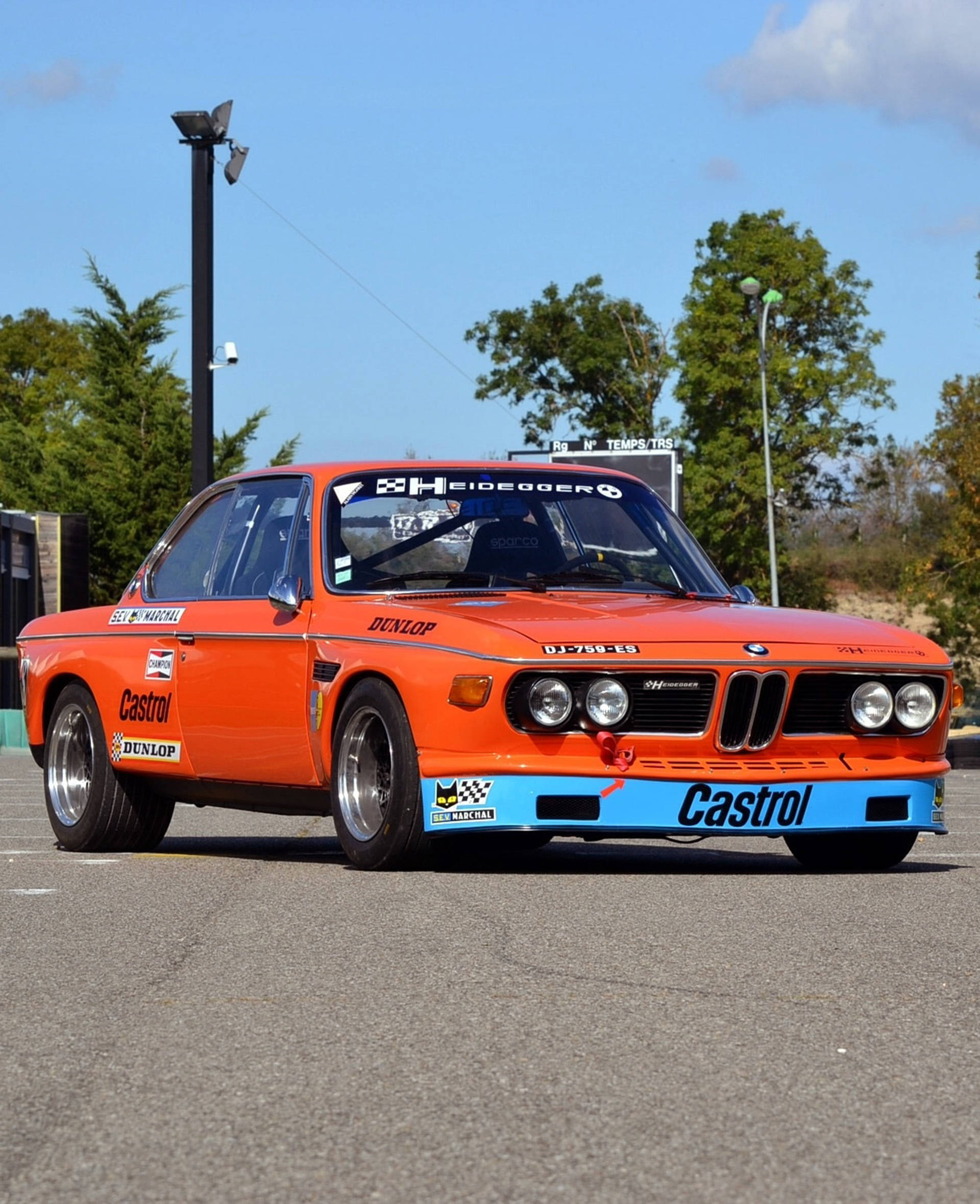 classic bmw 桌布 壁紙 — 一輛充滿活力的橙色復古賽車，帶有藍色點綴和Castrol貼紙，停在晴朗藍天下的瀝青路面上。