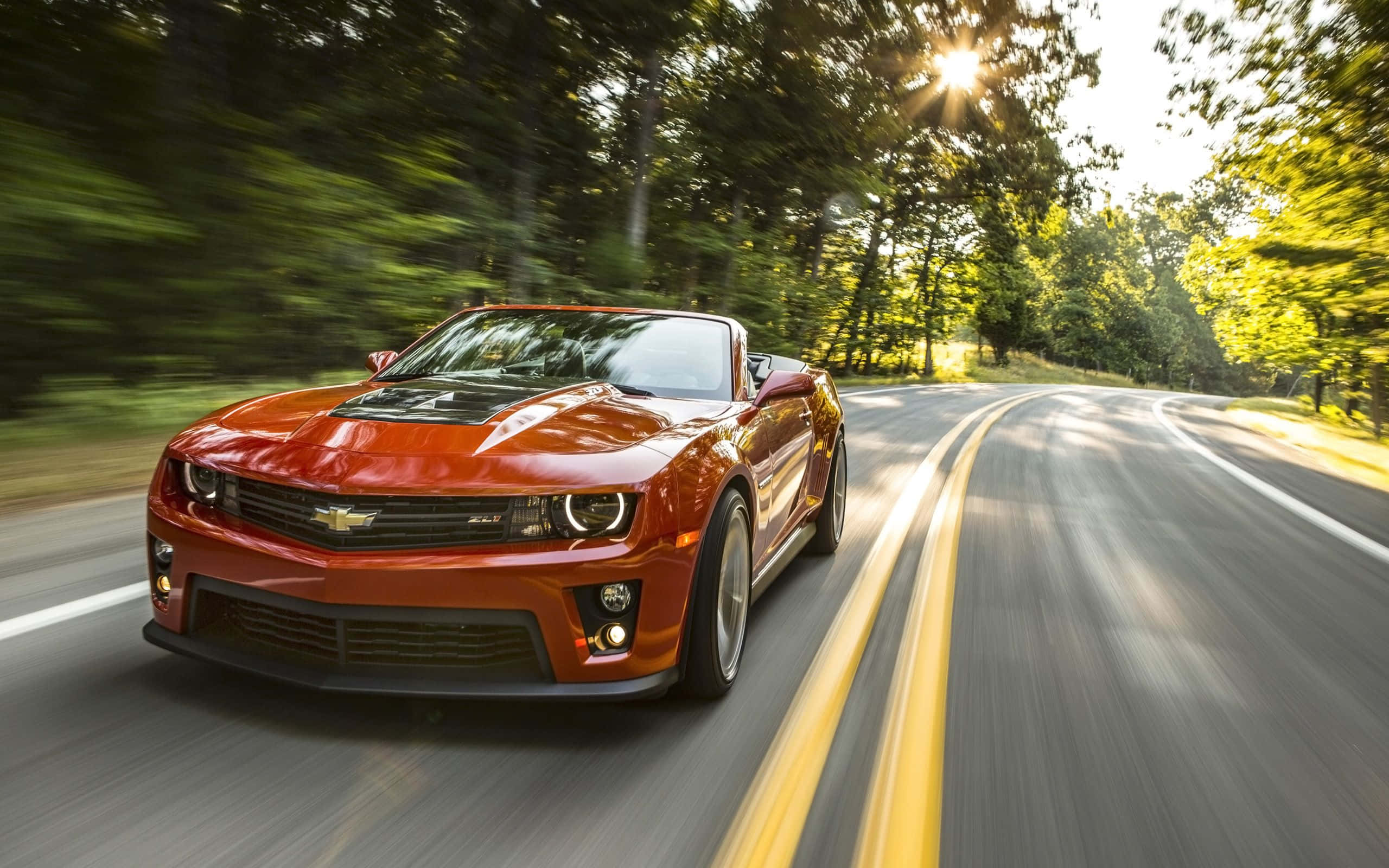 Cool Camaro 桌布 壁紙 — 一輛充滿活力的橙色敞篷跑車在蜿蜒的林間道路上疾馳，陽光穿過樹林照射下來。