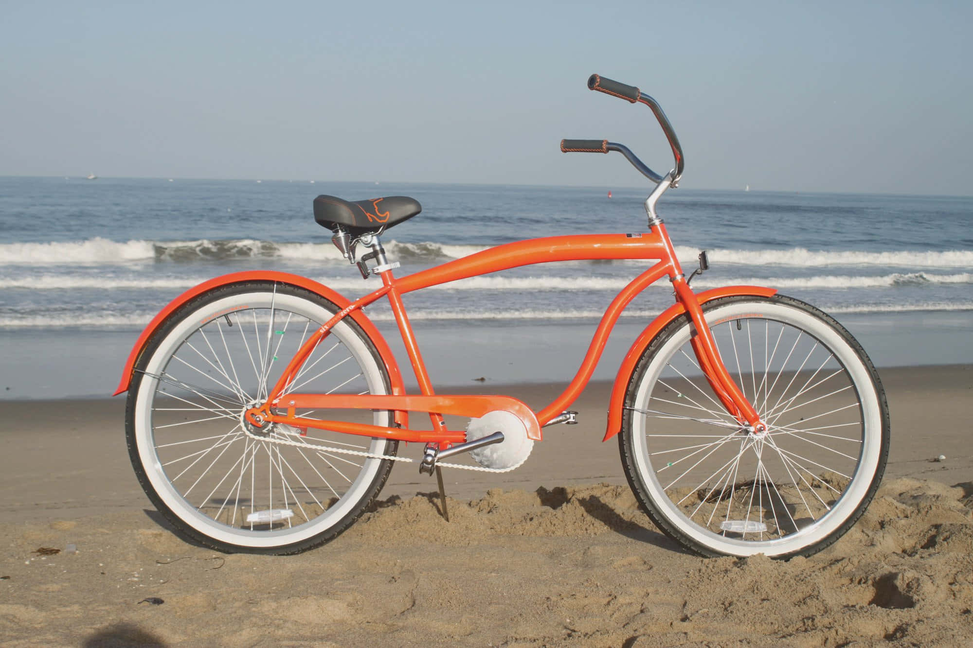 Beach Cruiser 桌布 壁紙 — 一輛充滿活力的橙色沙灘巡洋艦腳踏車停放在沙灘上，海浪在晴朗的天空下翻滾。