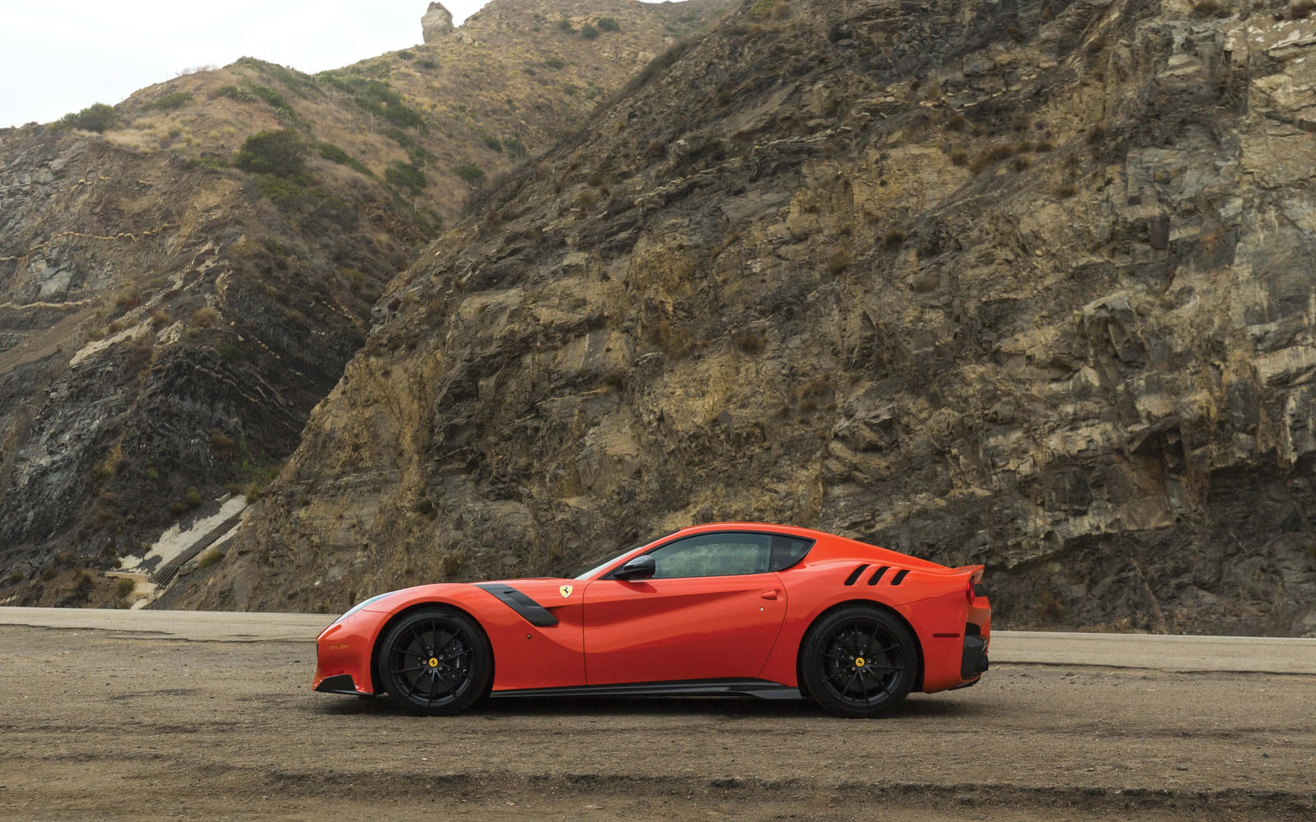 Ferrari F12 Berlinetta 桌布 壁紙 — 一輛充滿活力的橙色法拉利跑車停泊在塵土飛揚的路邊，背景是崎嶇的岩石峭壁。