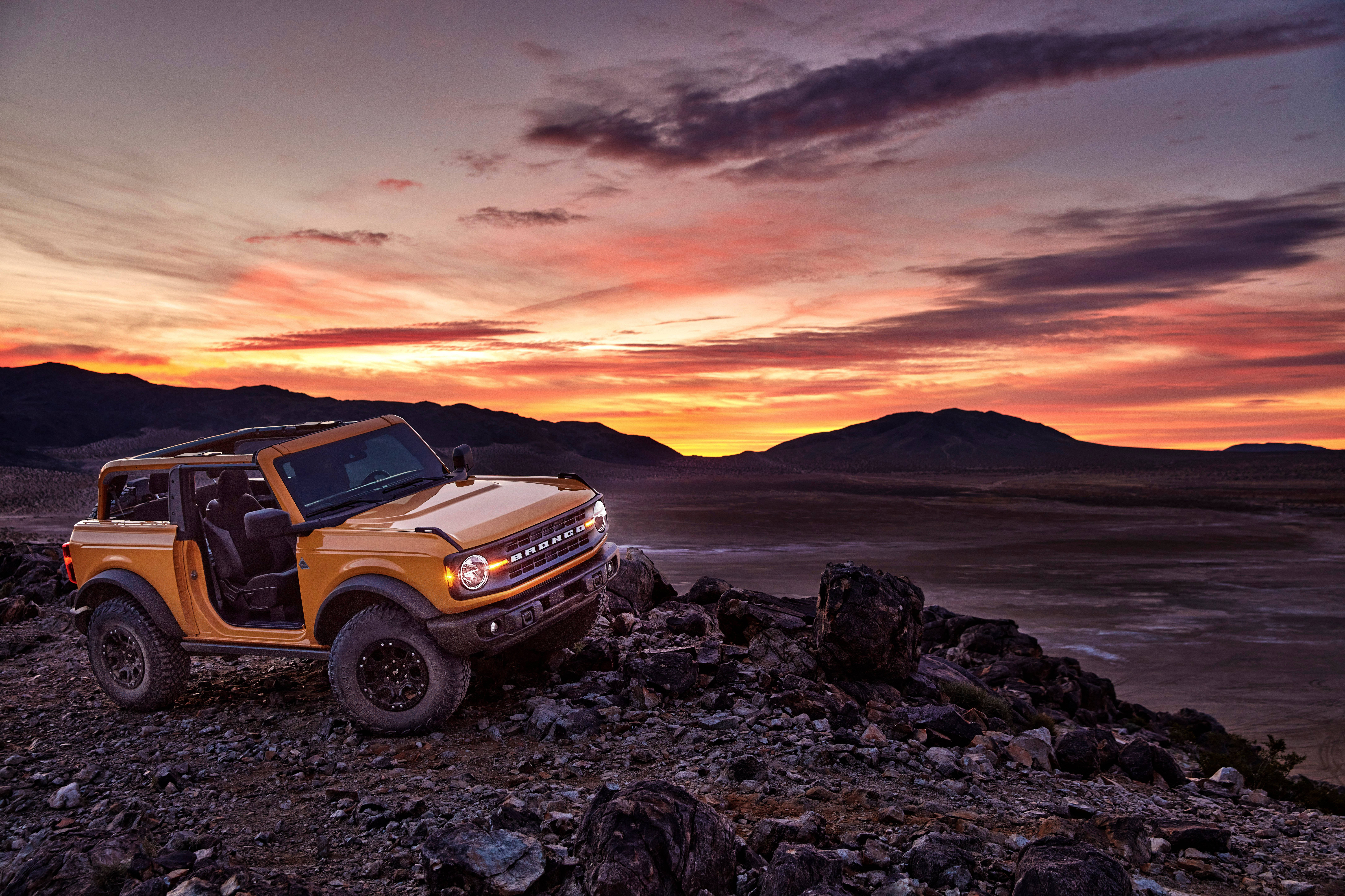 Ford Bronco 桌布 壁紙 — 一輛充滿活力的橙色福特Bronco在日落時分停在岩石地形上，遠山之上有火橙色和紫色的雲彩。