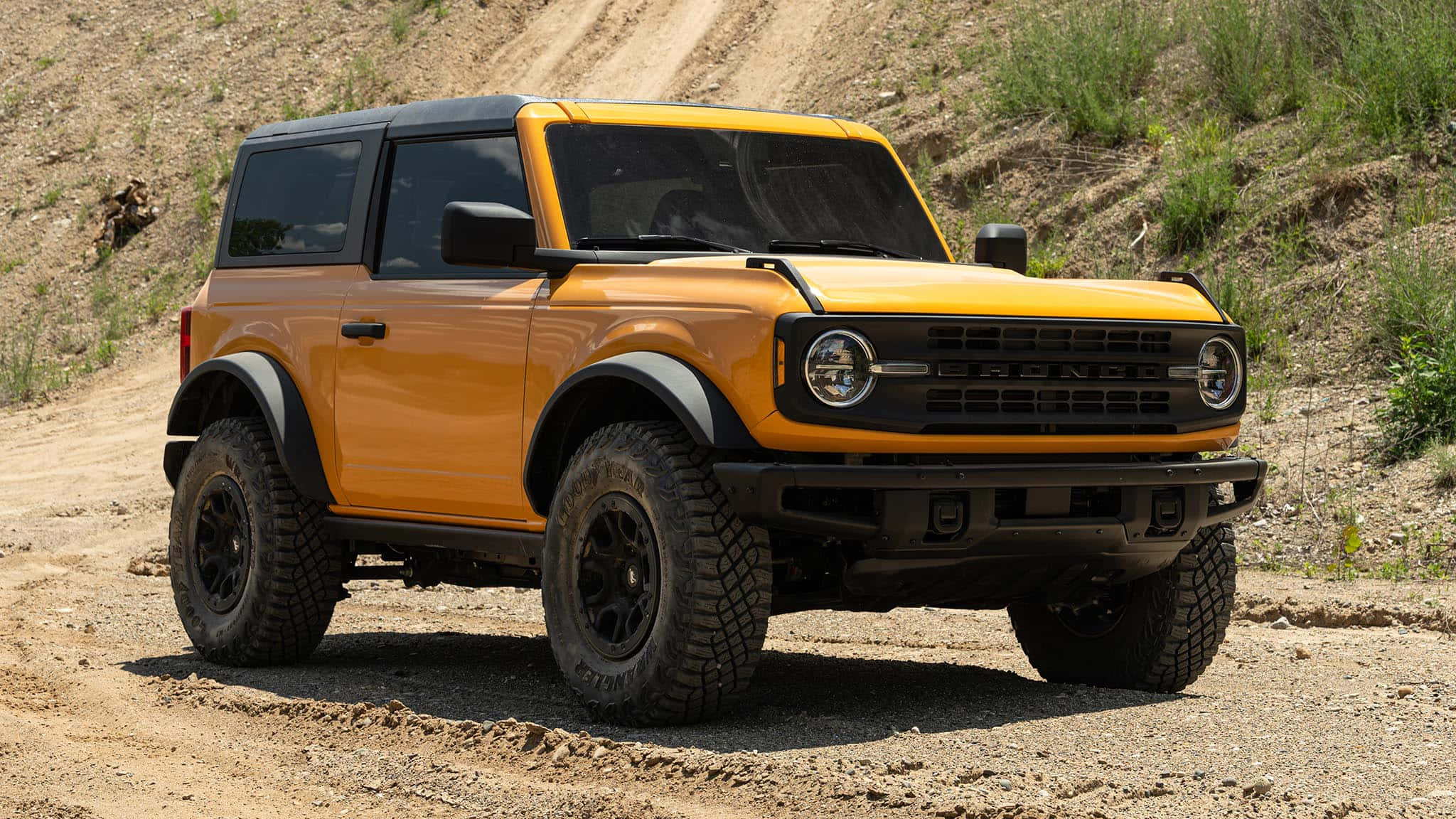 福特野馬越野車 桌布 壁紙 — 一輛充滿活力的橙色福特Bronco SUV，帶有黑色飾邊和堅固輪胎，在陽光明媚的泥土路上，以乾燥的草坡為背景，蓄勢待發。