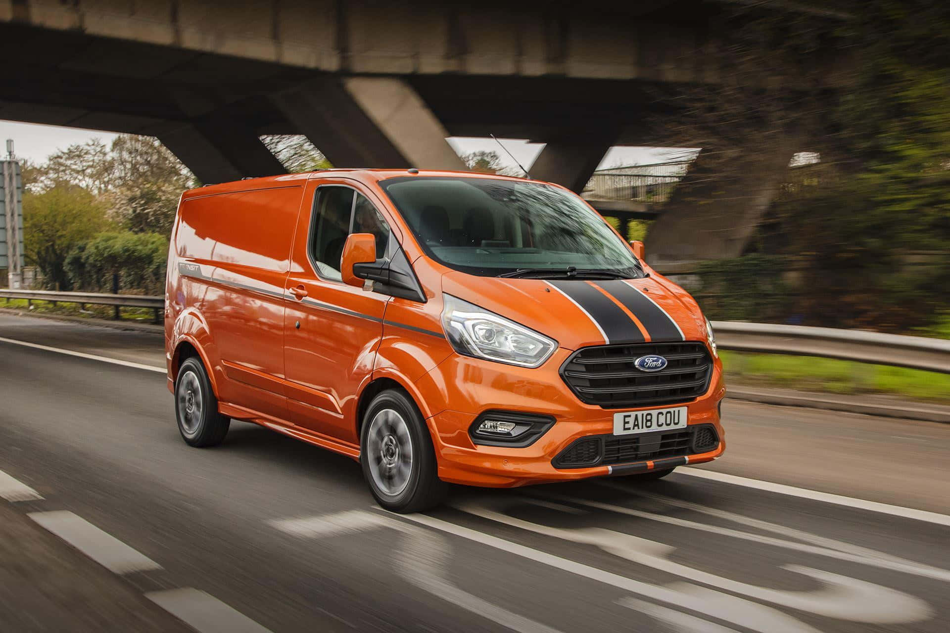 Ford Transit 桌布 壁紙 — 一輛充滿活力的橙色福特全順廂型車帶有黑色賽車條紋，在綠樹成蔭的道路上飛馳於混凝土高架橋下。