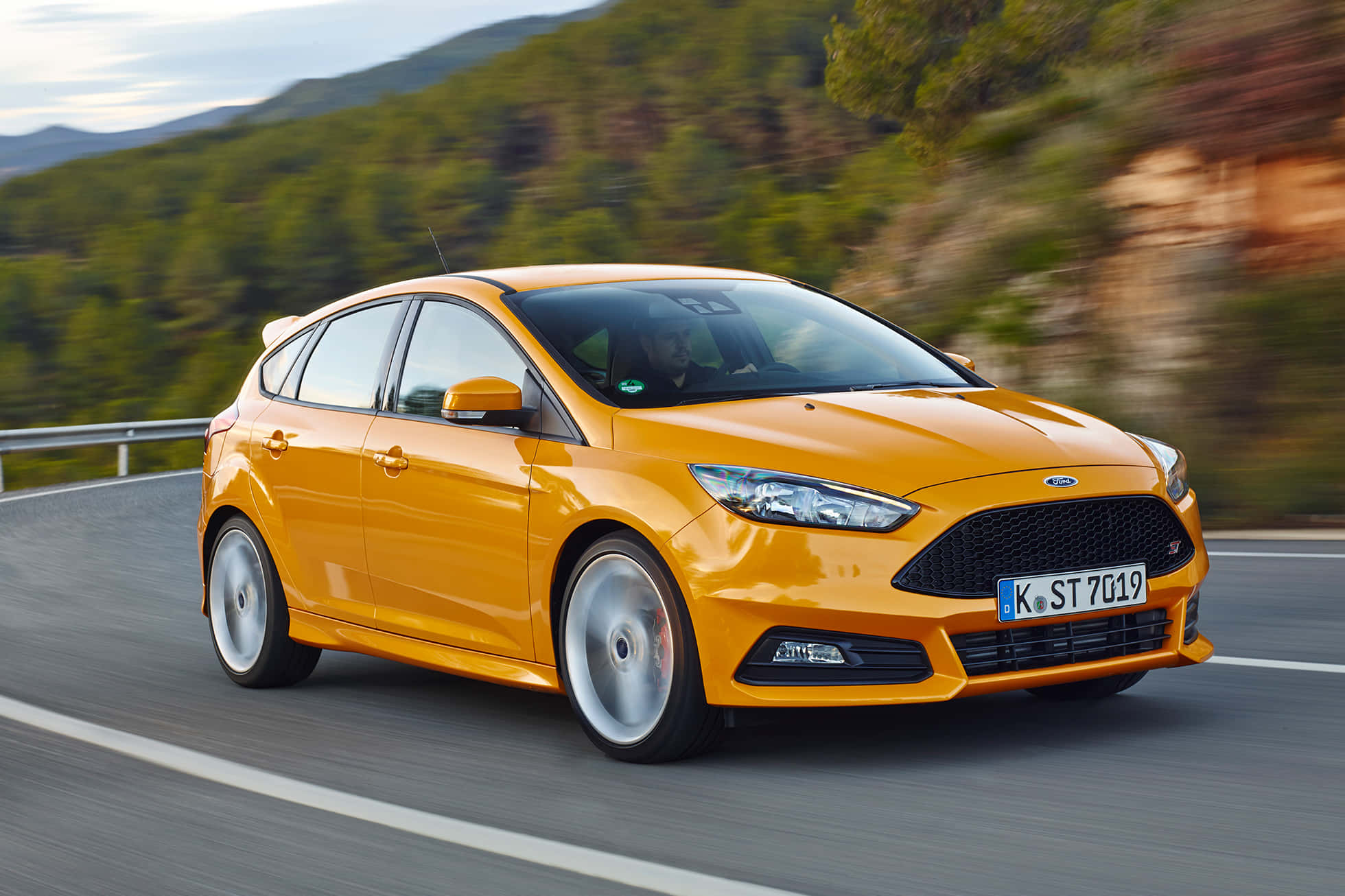 Ford Focus 桌布 壁紙 — 一輛充滿活力的橙色福特福克斯ST在蜿蜒的山路上奔馳，背景是模糊的綠色森林景色。