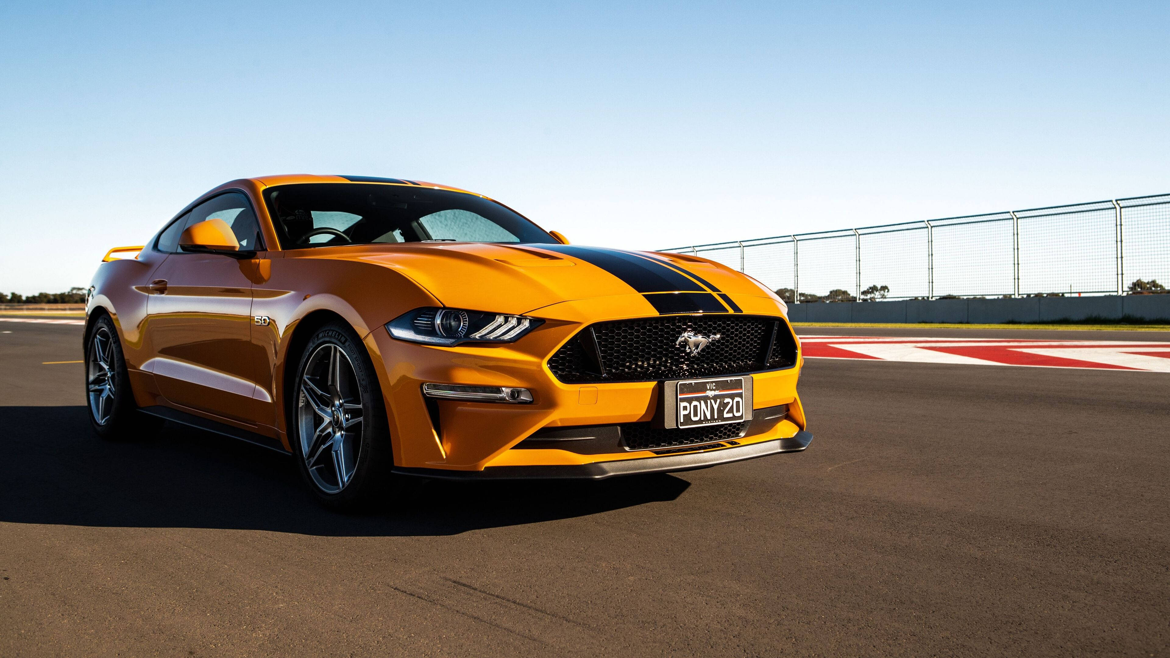 mustang HD 桌布 壁紙 — 一輛充滿活力的橙色福特野馬，帶有黑色賽車條紋，在晴朗的藍天下停在賽道上。