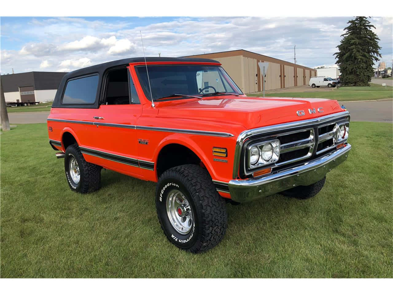 Gmc Jimmy 桌布 壁紙 — 一輛充滿活力的橙色經典GMC Jimmy SUV，帶有鍍鉻裝飾和升高懸掛，自豪地停在綠色草坪上，天空部分多雲。