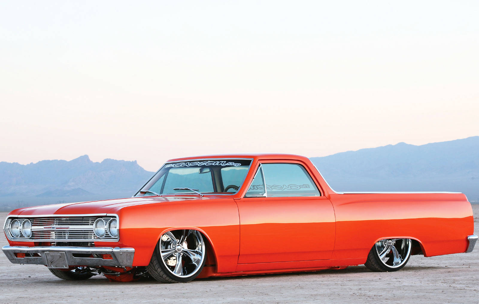 Lowrider 桌布 壁紙 — 一輛充滿活力的橙色經典低底盤皮卡車在柔和的暮光下，映襯著遠處的山脈閃閃發光。