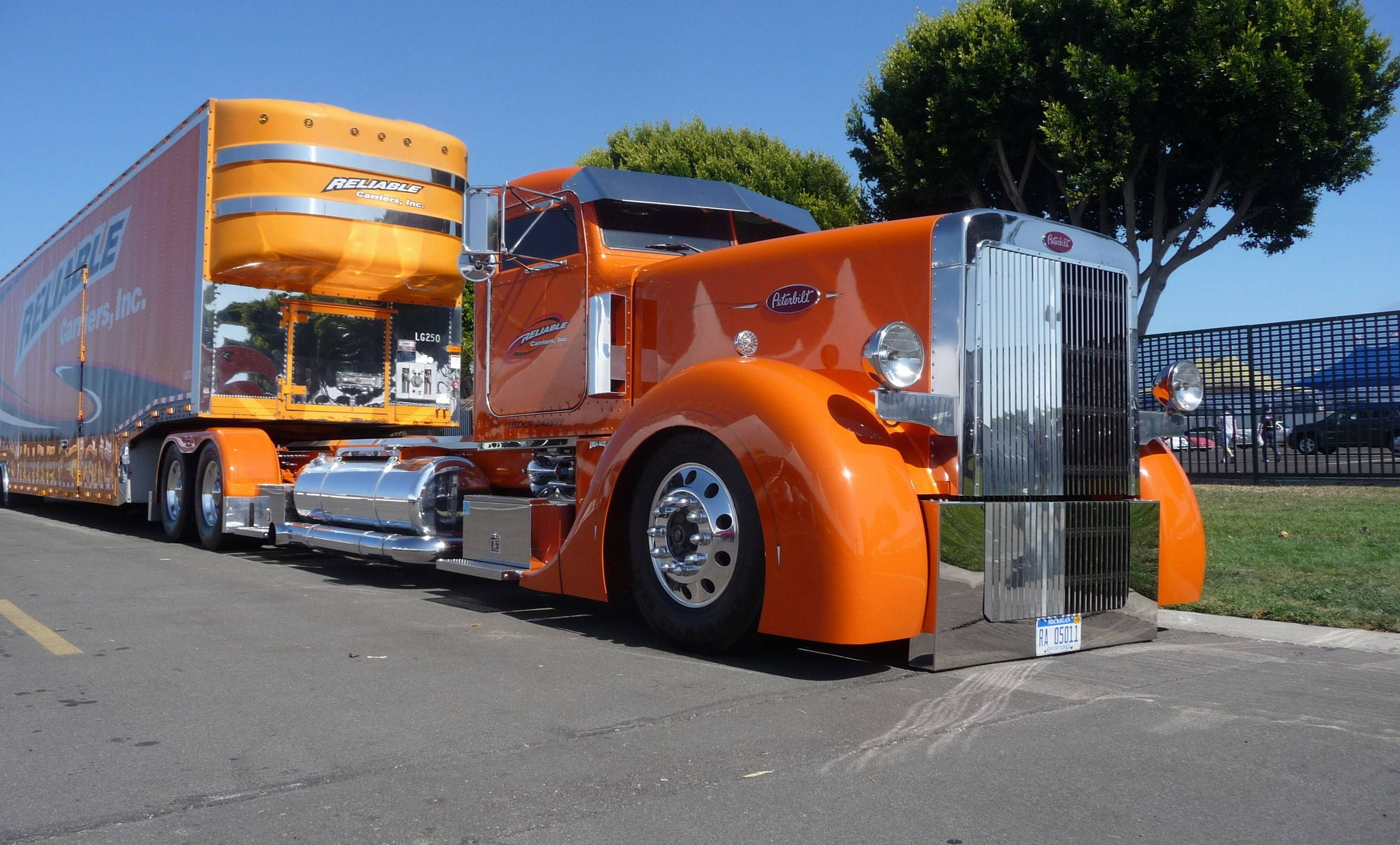 Peterbilt 桌布 壁紙 — 一輛充滿活力的橙色經典半掛卡車，帶有鍍鉻裝飾和匹配的拖車，停在晴朗藍天下的瀝青路面上。