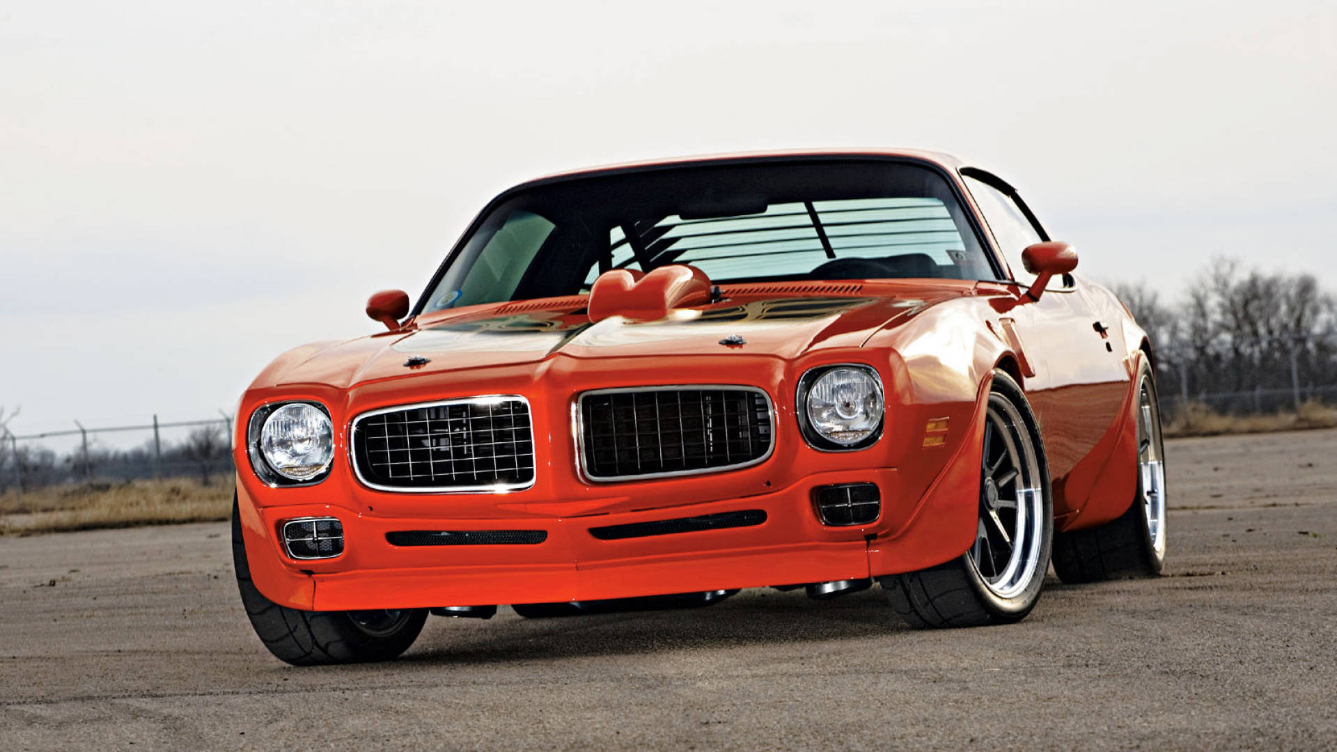 Pontiac 桌布 壁紙 — 一輛充滿活力的橙色經典肌肉車，配備定制寬體套件和鍍鉻格柵，停放在開闊的瀝青地上，天空多雲。