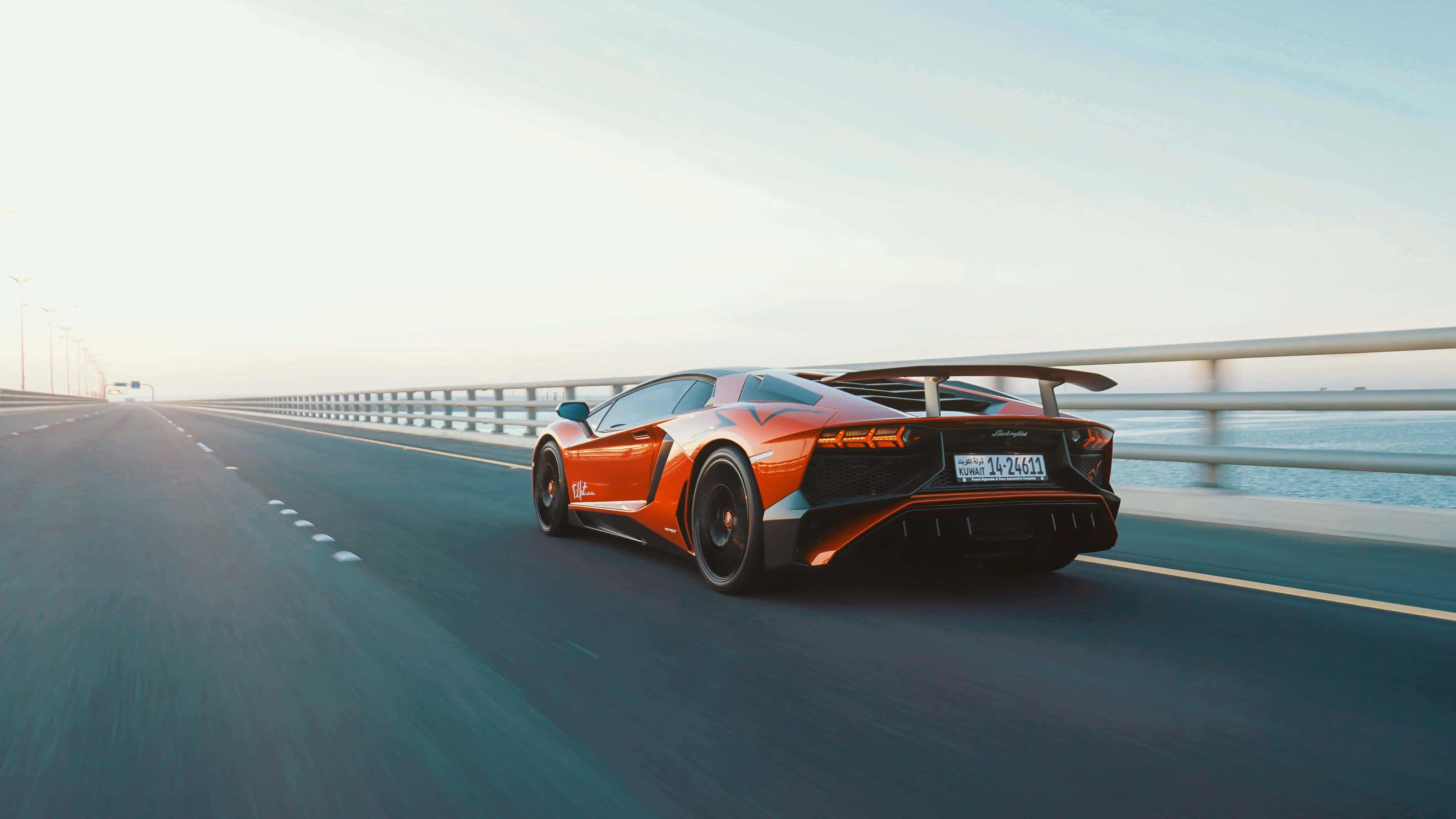蘭博基尼 桌布 壁紙 — 一輛充滿活力的橙色藍寶堅尼 Aventador 沿著海岸高速公路疾馳，帶有模糊的動感和平靜的海洋背景。