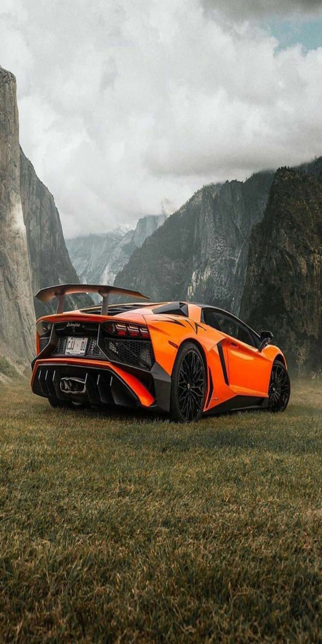 Pixel 3 Lamborghini 桌布 壁紙 — 一輛充滿活力的橙色藍寶堅尼 Aventador 停泊在草地地形上，背景有高聳的岩石懸崖和陰天。