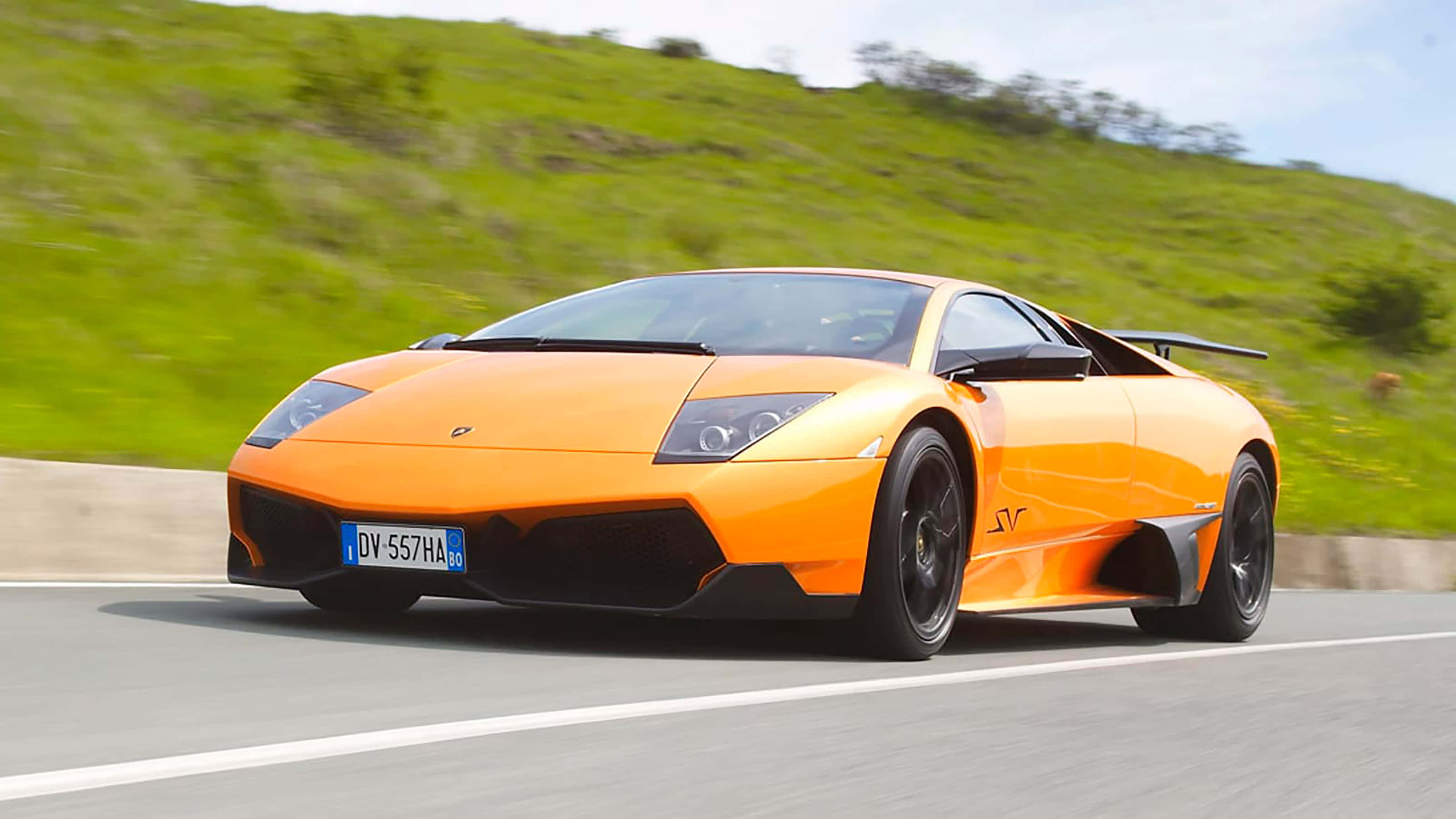 Cool Lamborghinis 桌布 壁紙 — 一輛充滿活力的橙色藍寶堅尼 Murciélago SV 在蜿蜒的道路上奔馳，背景是模糊的綠色山丘。