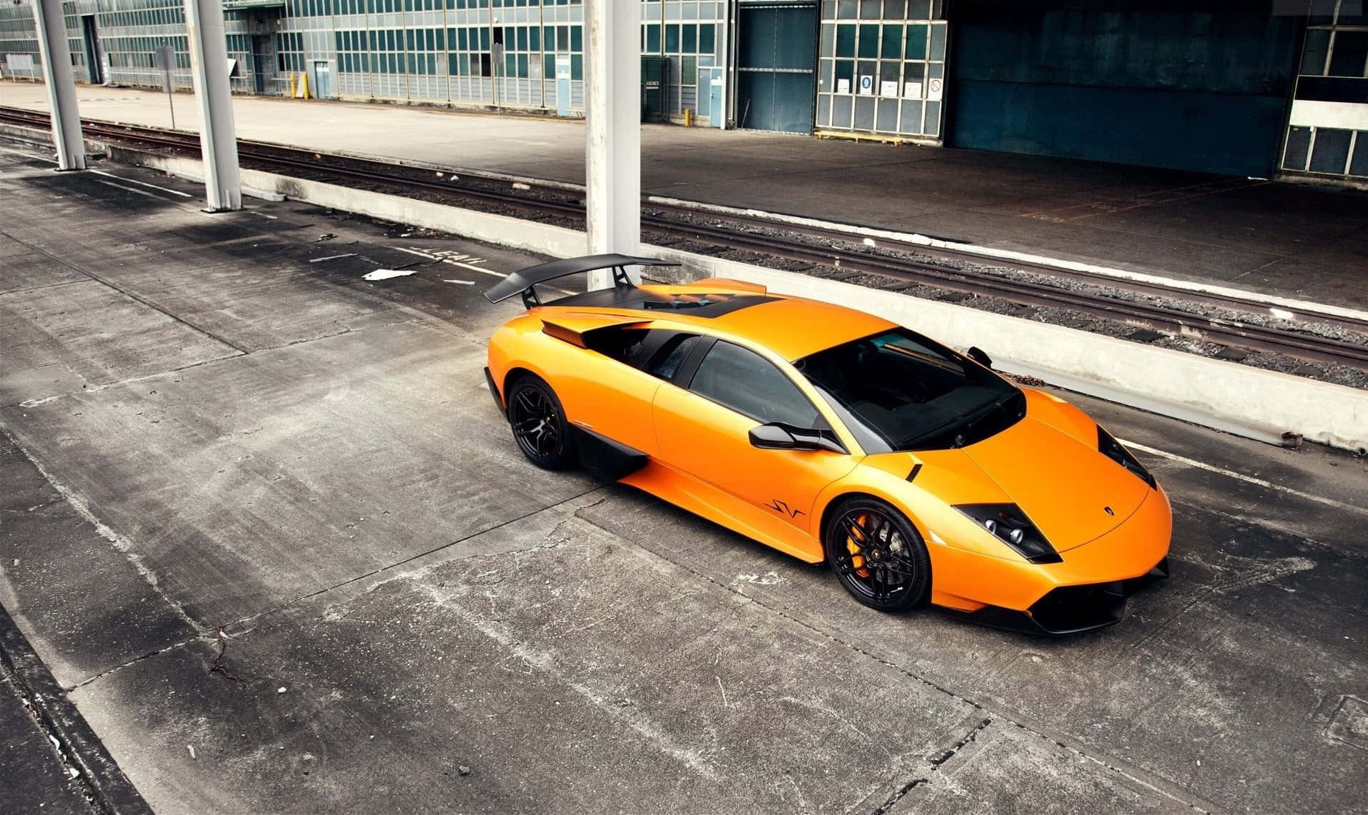Lamborghini Murciélago 桌布 壁紙 — 一輛充滿活力的橙色藍寶堅尼 Murciélago 停放在工業建築物下方、火車軌道旁的破裂混凝土上。