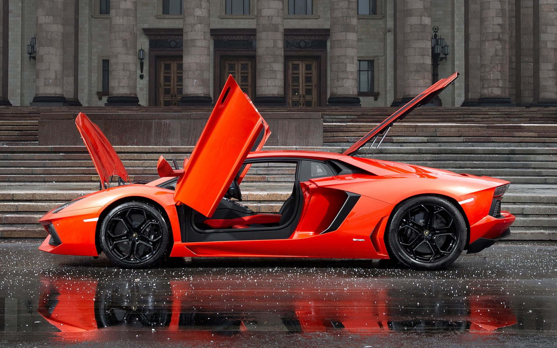 Cool Car 桌布 壁紙 — 一輛充滿活力的橙色藍寶堅尼Aventador，剪刀門打開，停在潮濕的路面上，其大膽的造型映襯著宏偉的石建築。
