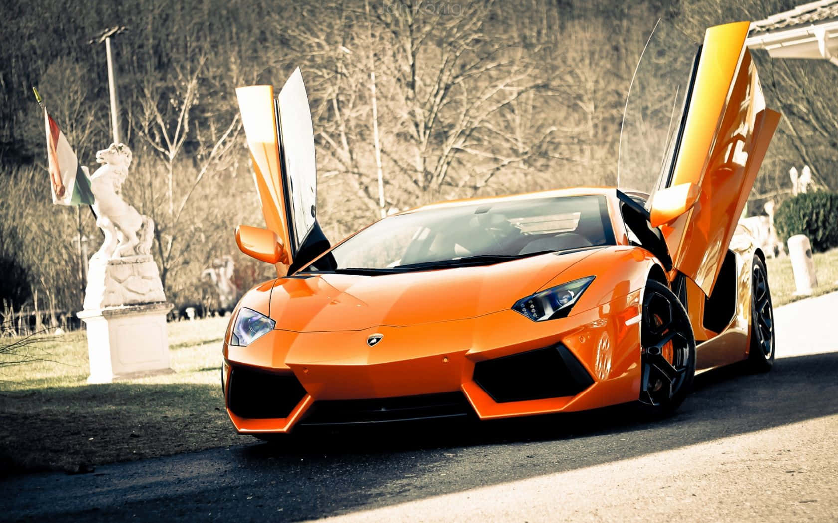 跑車 桌布 壁紙 — 一輛充滿活力的橙色藍寶堅尼Aventador，剪刀門打開，停泊在車道上，旁邊有一座白色大理石雕像。