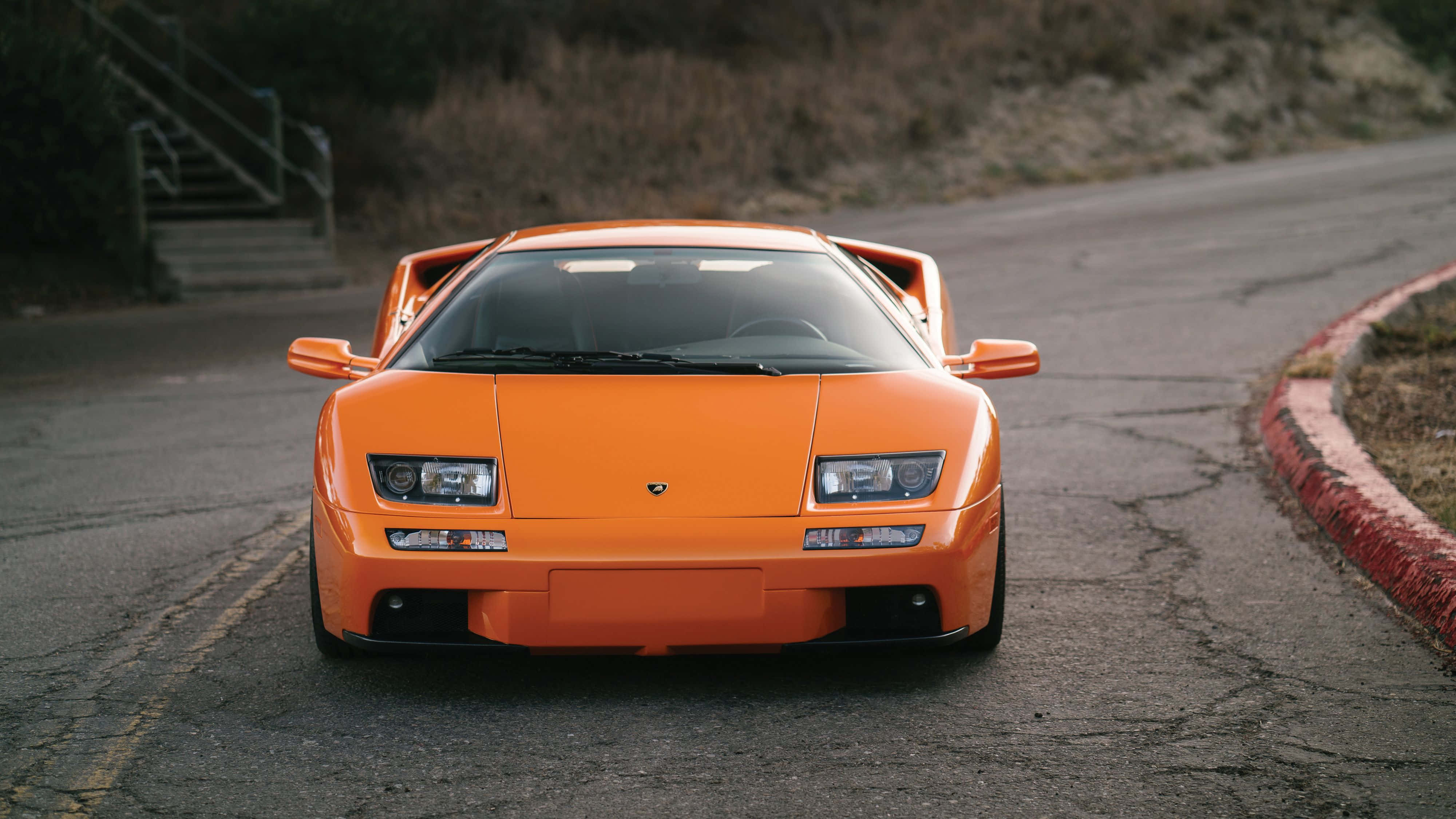 Lamborghini Diablo 桌布 壁紙 — 一輛充滿活力的橙色藍寶堅尼Diablo跑車，在蜿蜒的柏油路上向前行駛，路面有裂縫和乾草。