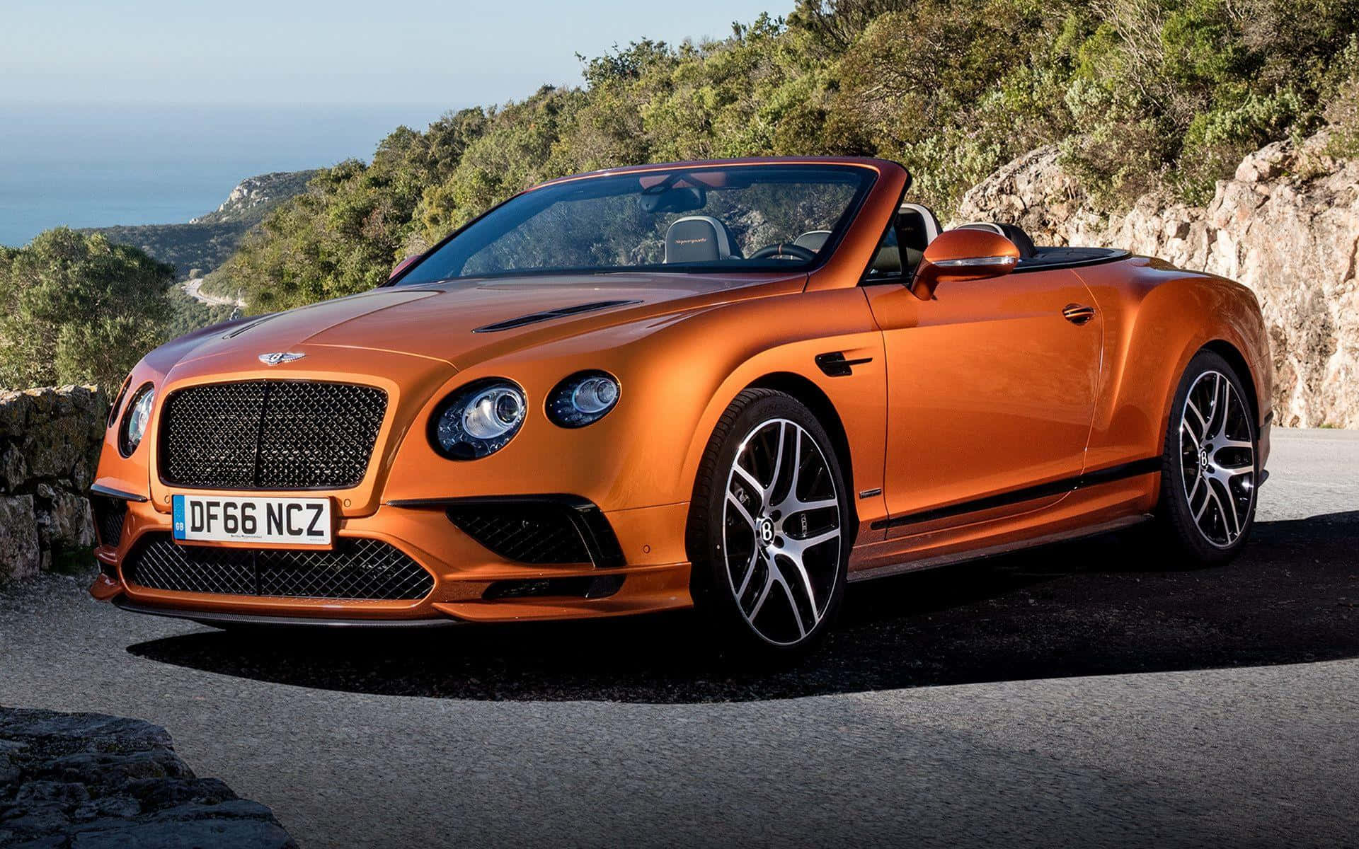 bentley HD 桌布 壁紙 — 一輛充滿活力的橙色賓利敞篷車停泊在沿海公路上，背景是鬱鬱蔥蔥的綠色山丘和海洋。