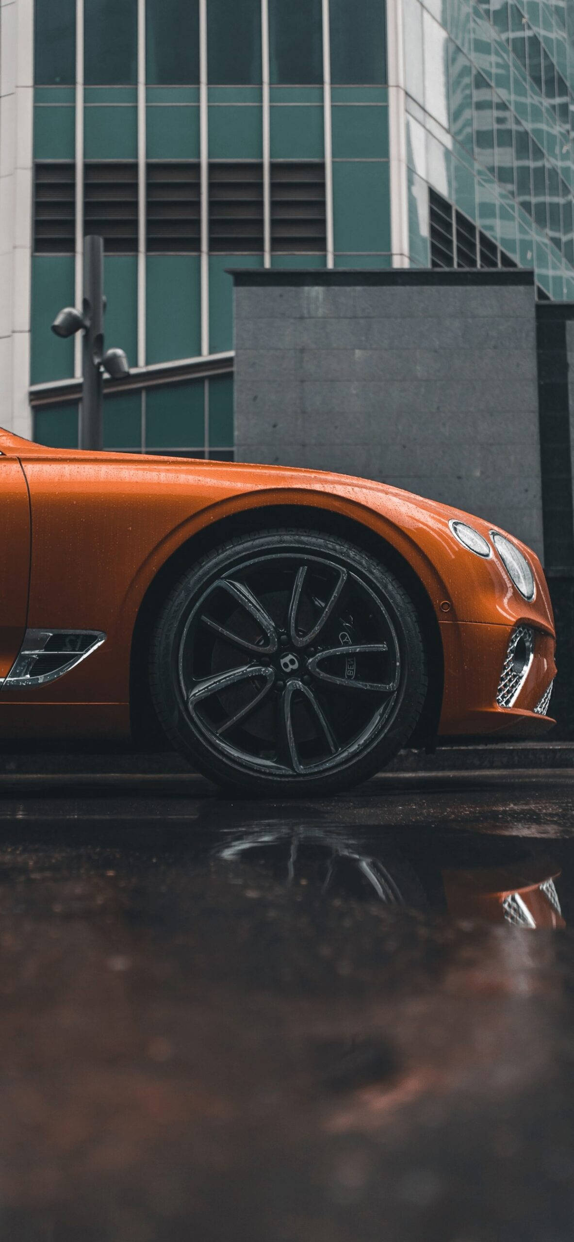 bentley iPhone 桌布 壁紙 — 一輛充滿活力的橙色賓利跑車在濕漉漉的路面上閃閃發光，背後是現代化的玻璃建築高聳入雲。