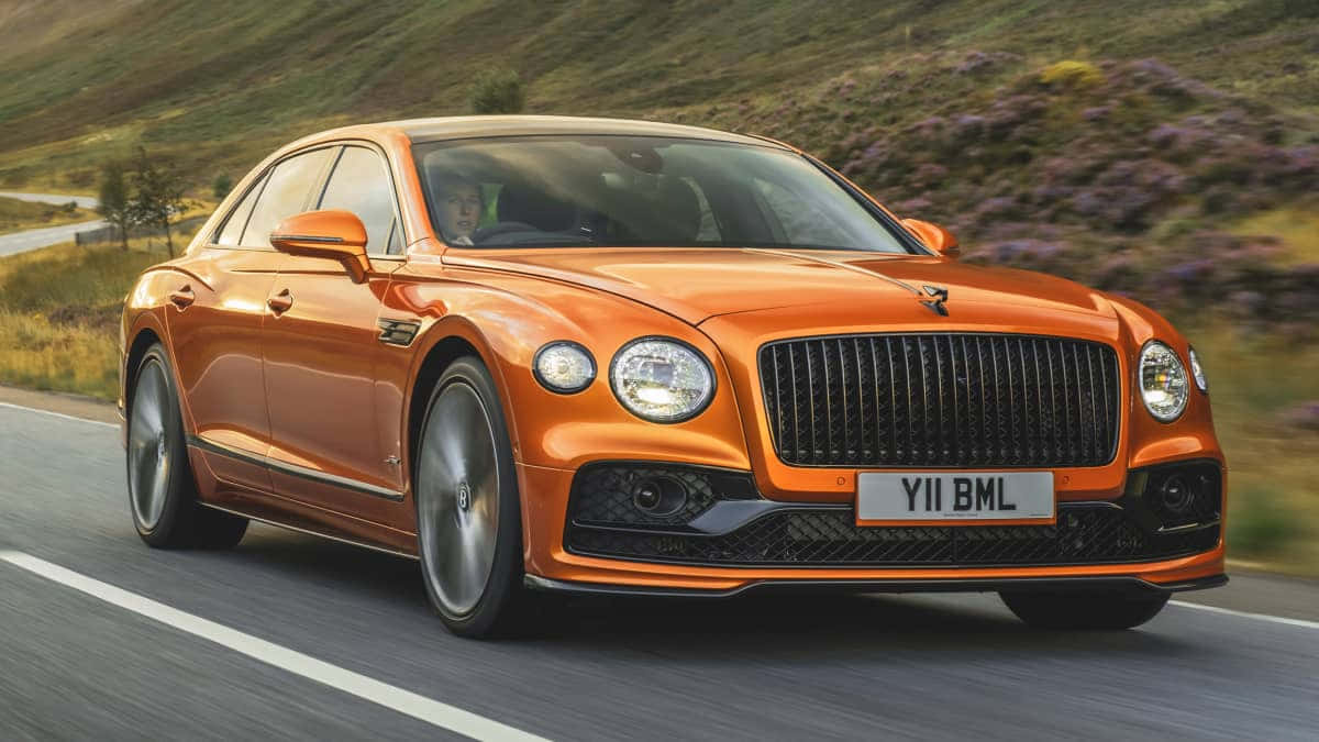 Bentley Flying Spur 桌布 壁紙 — 一輛充滿活力的橙色賓利飛馳在蜿蜒的山路上，背景是模糊的紫色石楠花。