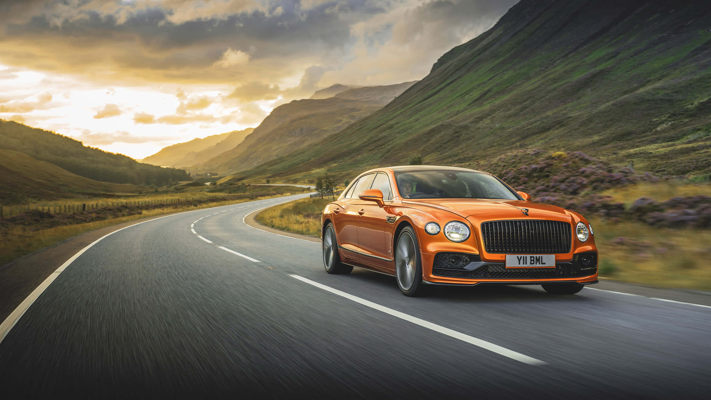 Bentley Mulsanne 桌布 壁紙 — 一輛充滿活力的橙色賓利飛馳在黃金時段沿著蜿蜒的山路飛馳，頭頂上有戲劇性的雲彩。