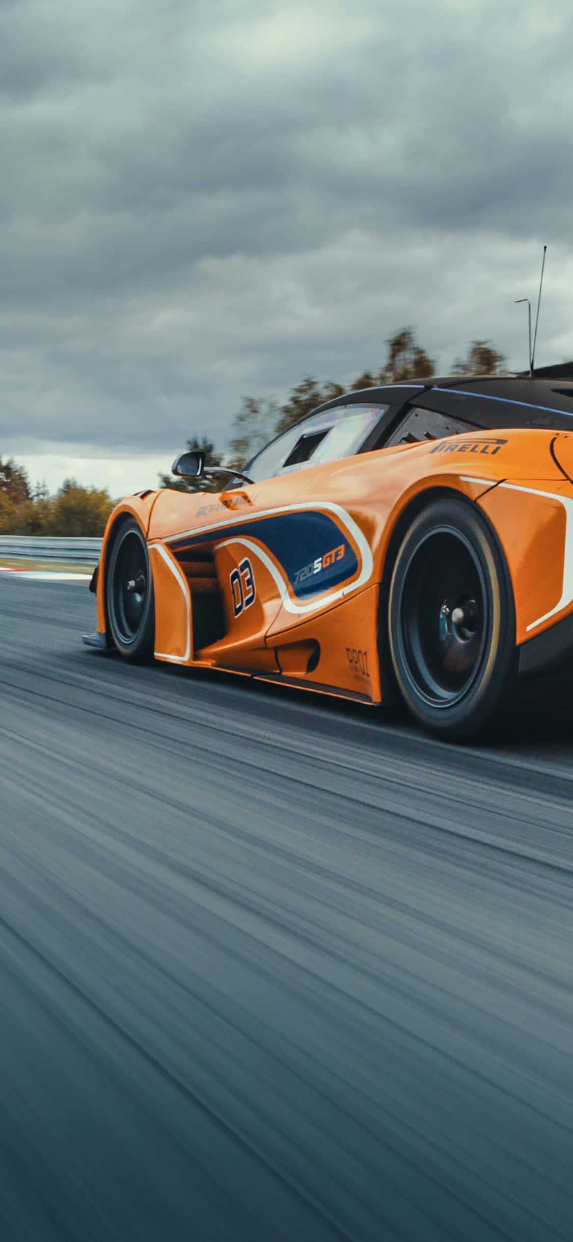 iPhone Xs Mclaren 720s 桌布 壁紙 — 一輛充滿活力的橙色賽車在潮濕的賽道上奔馳，天空多雲，帶有模糊的動態線條。