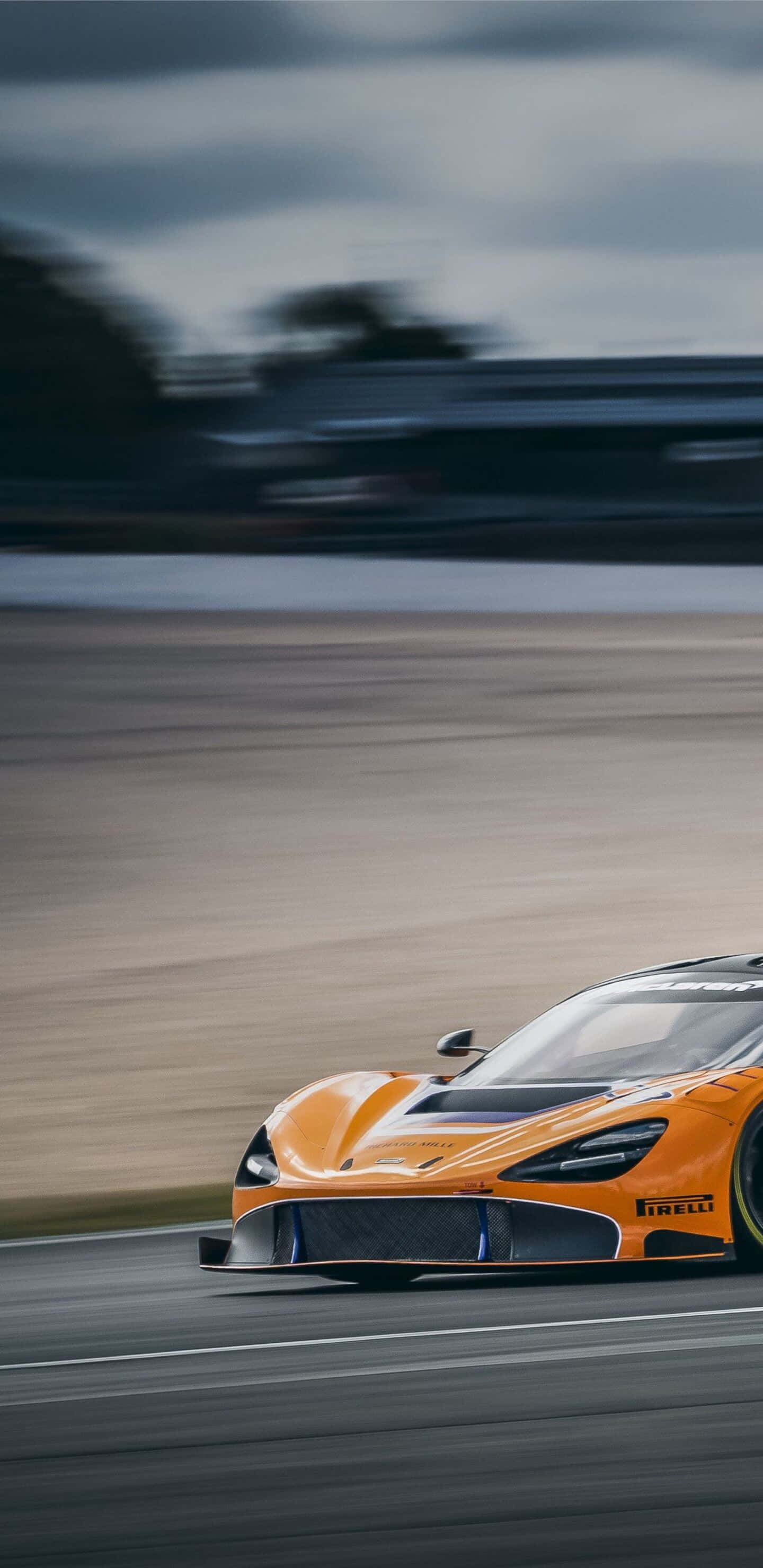 Pixel 3xl Mclaren 720s 桌布 壁紙 — 一輛充滿活力的橙色賽車在陰天下的賽道上疾馳而過，運動條紋強調了速度。
