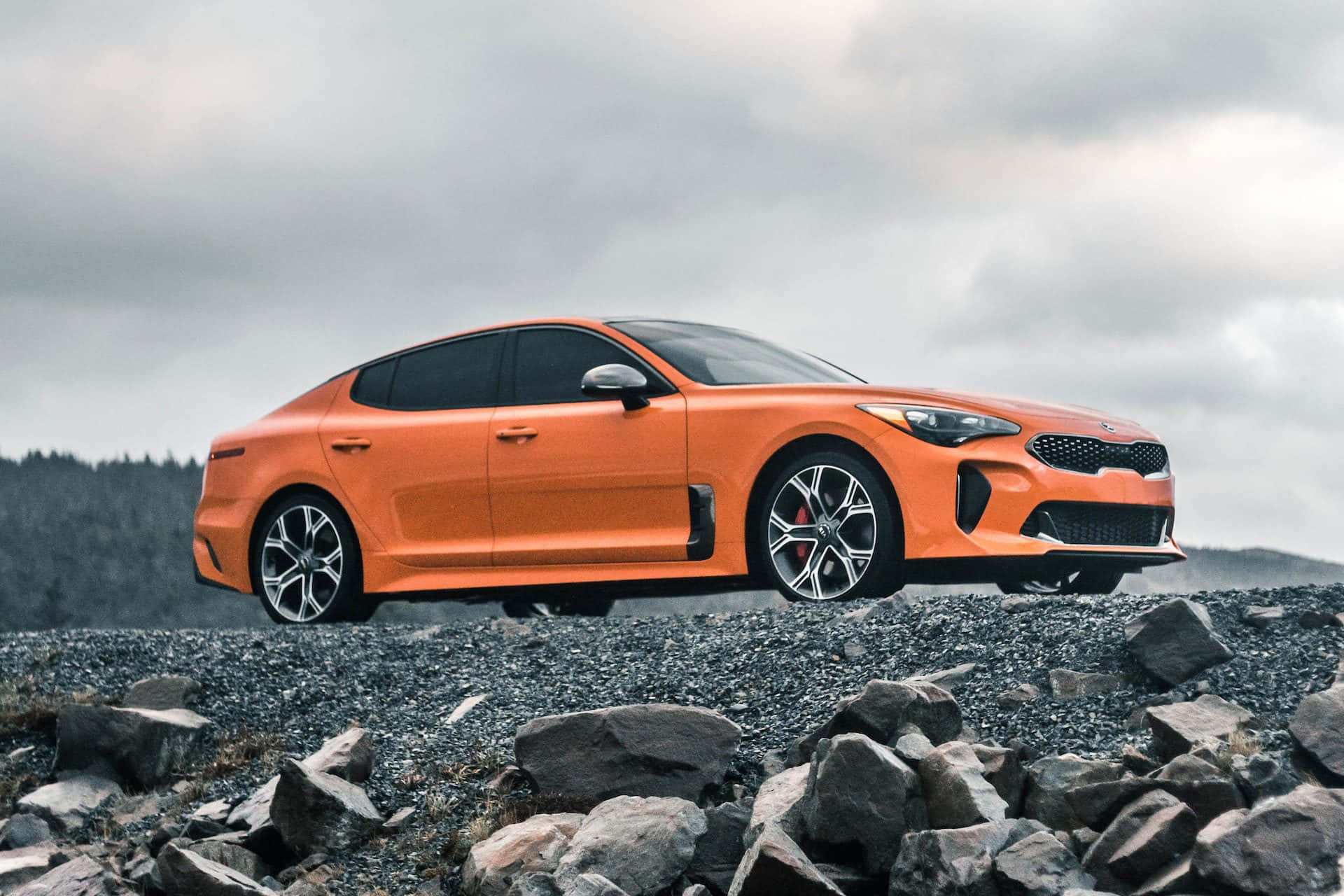Kia Stinger 桌布 壁紙 — 一輛充滿活力的橙色跑車停泊在岩石山坡上，天空多雲。
