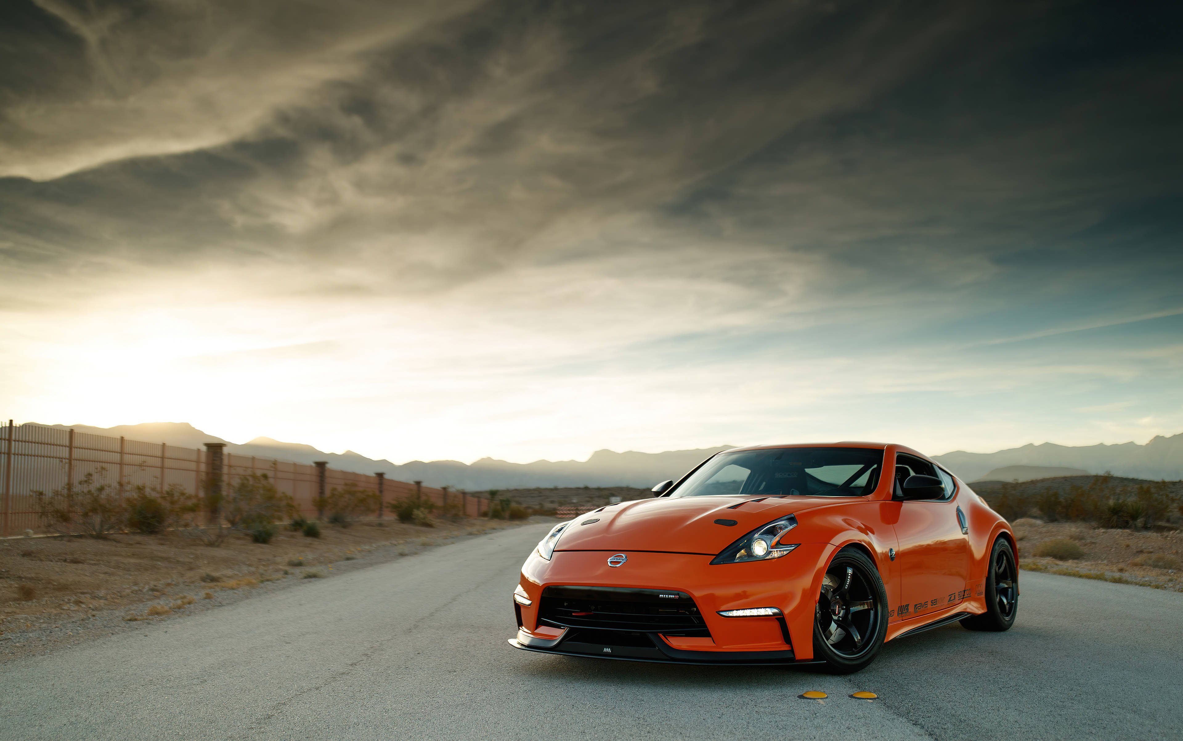 Nissan 370z 桌布 壁紙 — 一輛充滿活力的橙色跑車停泊在沙漠公路上，背景是戲劇性的多雲日落天空。