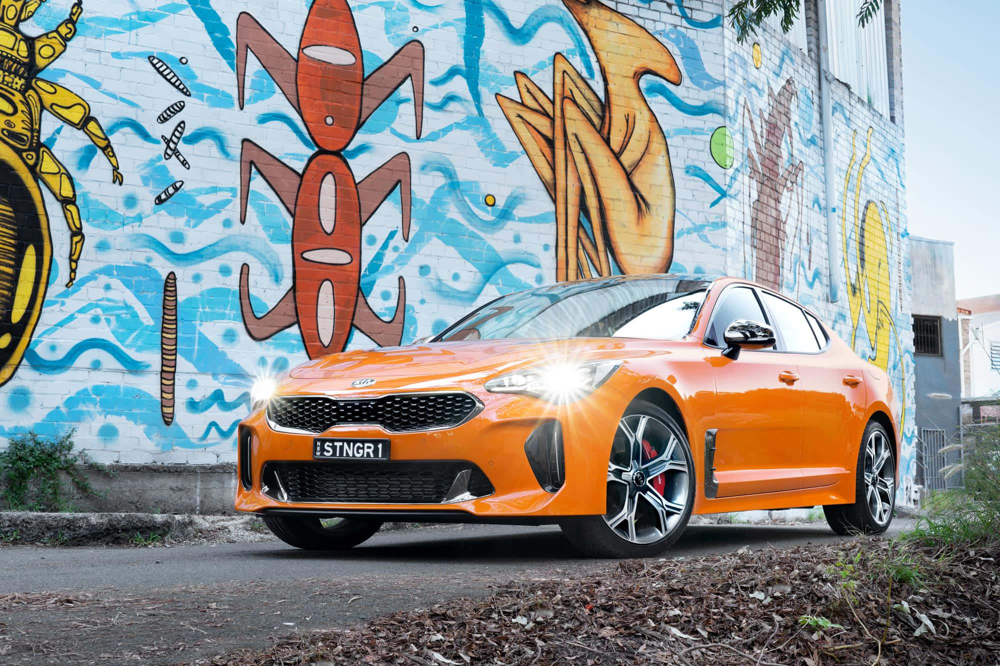 Kia Stinger 桌布 壁紙 — 一輛充滿活力的橙色跑車在日光下閃閃發光，停在一幅色彩繽紛的塗鴉城市牆前。