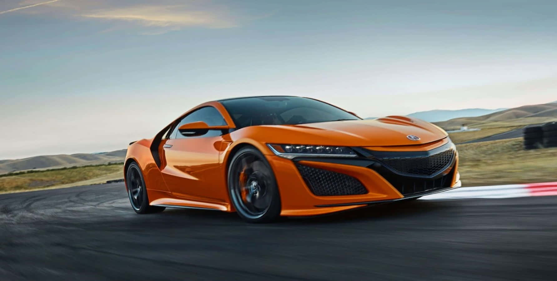 Acura 桌布 壁紙 — 一輛充滿活力的橙色跑車在蜿蜒的賽道上疾馳，背景是朦朧的天空和起伏的山丘，呈現出模糊的動感。