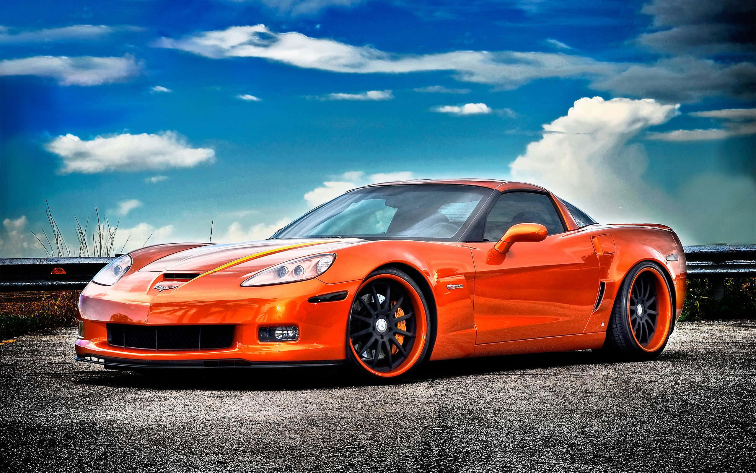 Chevrolet Corvette Z06 桌布 壁紙 — 一輛充滿活力的橙色跑車搭配黑色輪圈，停在路邊，背景是戲劇性的藍天和蓬鬆的白雲。