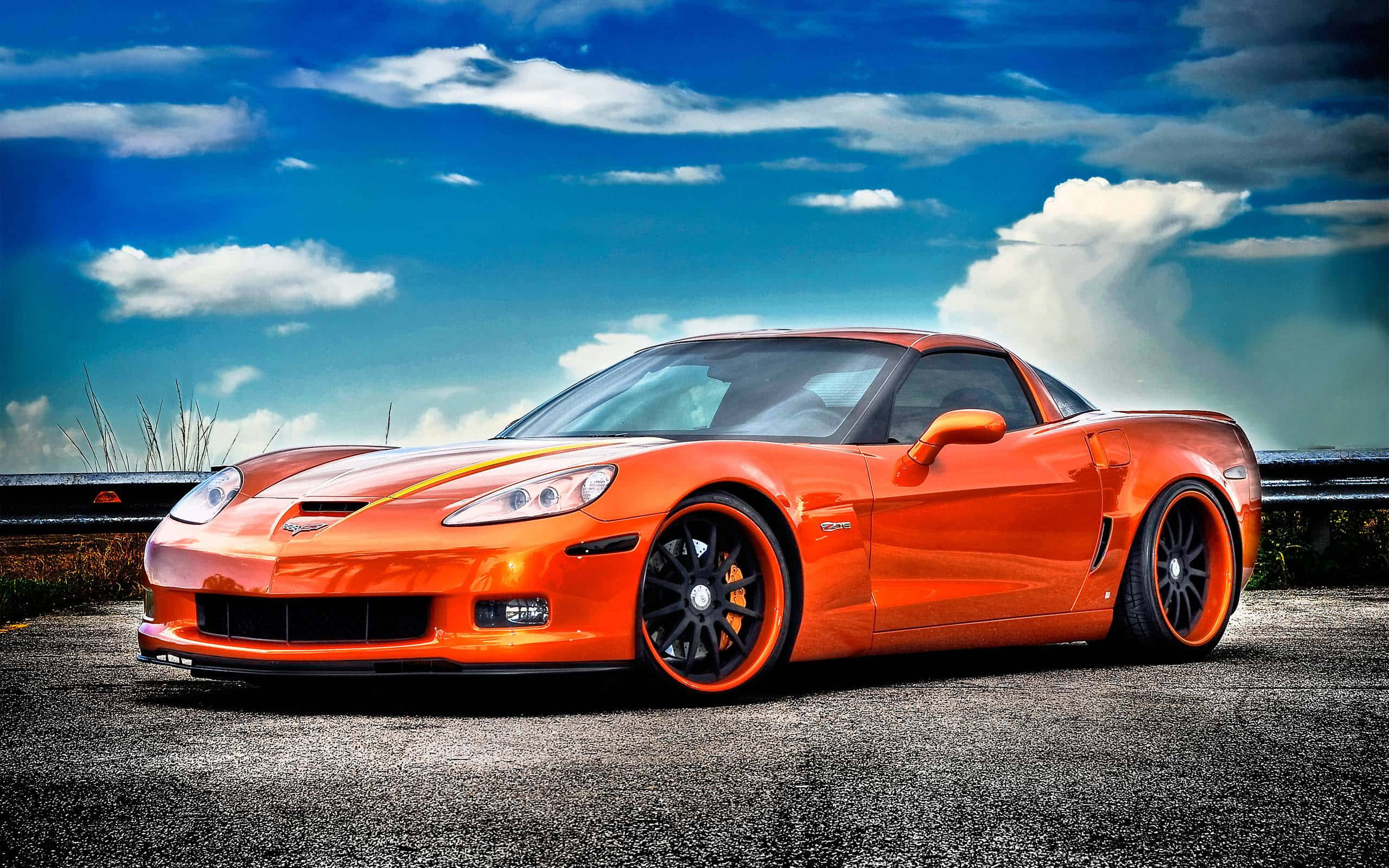 corvette 桌布 壁紙 — 一輛充滿活力的橙色跑車配黑色輪圈，停在路邊，天空湛藍，白雲朵朵。