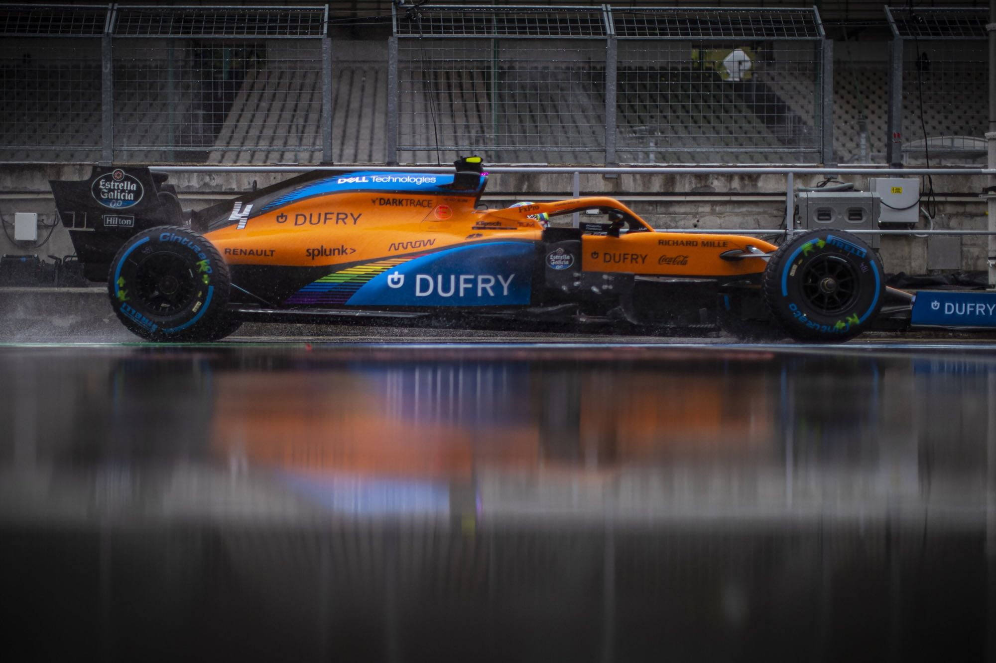 F1 桌布 壁紙 — 一輛充滿活力的橙藍色一級方程式賽車在潮濕賽道的水坑中飛馳，倒映其大膽塗裝於陰天之下。