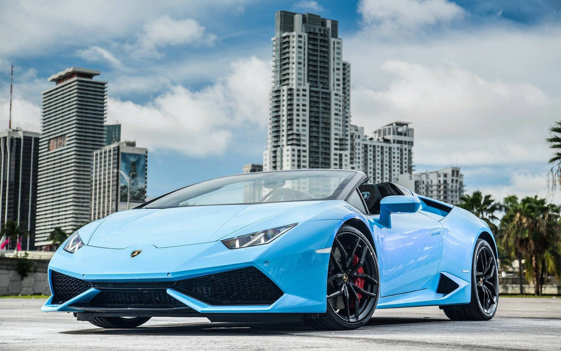 藍色 桌布 壁紙 — 一輛充滿活力的淺藍色藍寶堅尼 Huracán Spyder 停泊在現代摩天大樓和棕櫚樹林立的城市街道上，天空部分多雲。