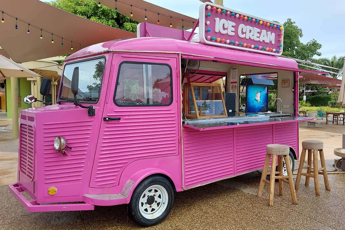 Ice Cream Truck 桌布 壁紙 — 一輛充滿活力的粉紅色復古冰淇淋車停放在帶有燈串和木凳的遮陽篷下。