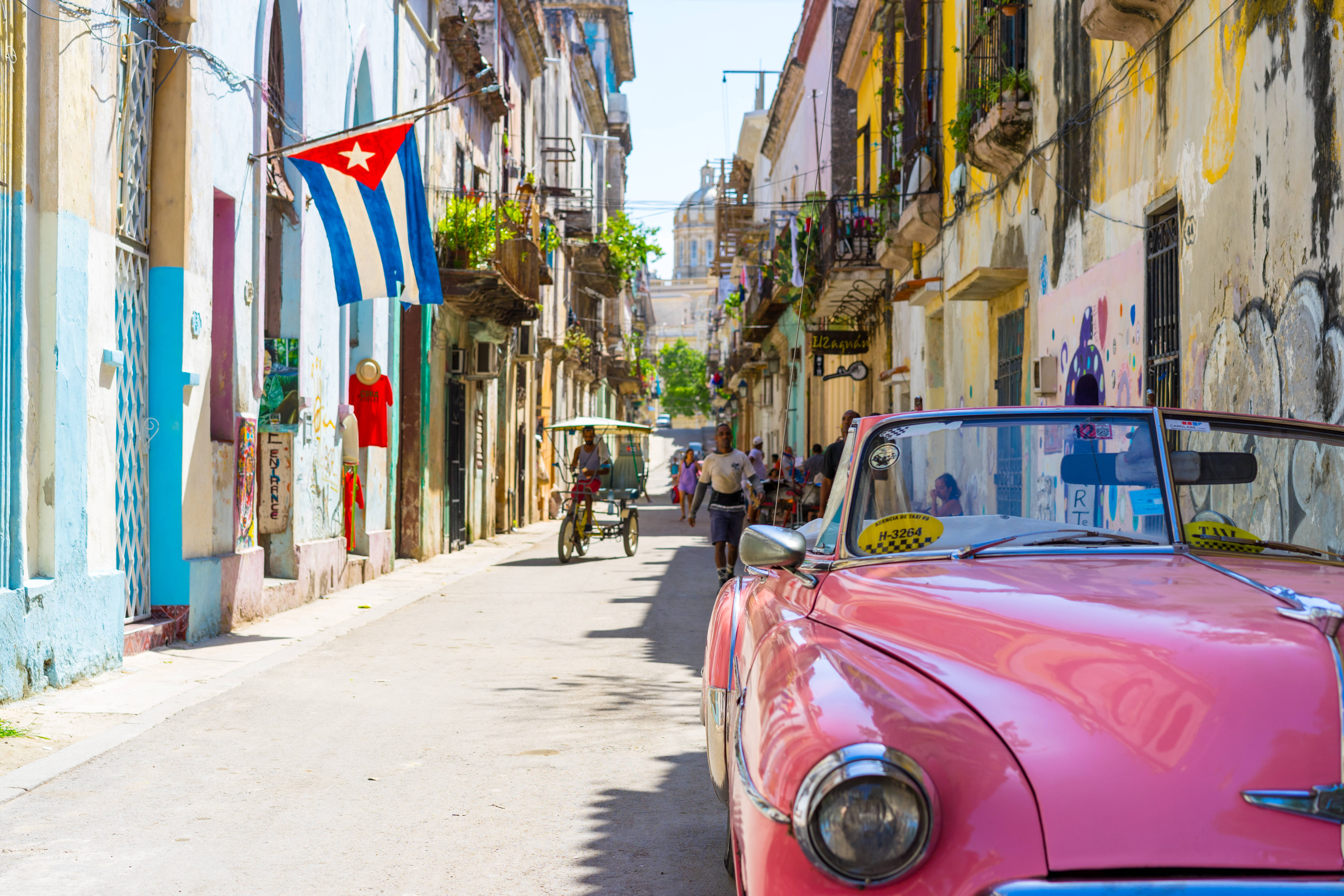 Cuba 桌布 壁紙 — 一輛充滿活力的粉紅色復古車停在陽光明媚的古巴街道上，街道兩旁是色彩繽紛的殖民建築和飄揚的國旗。